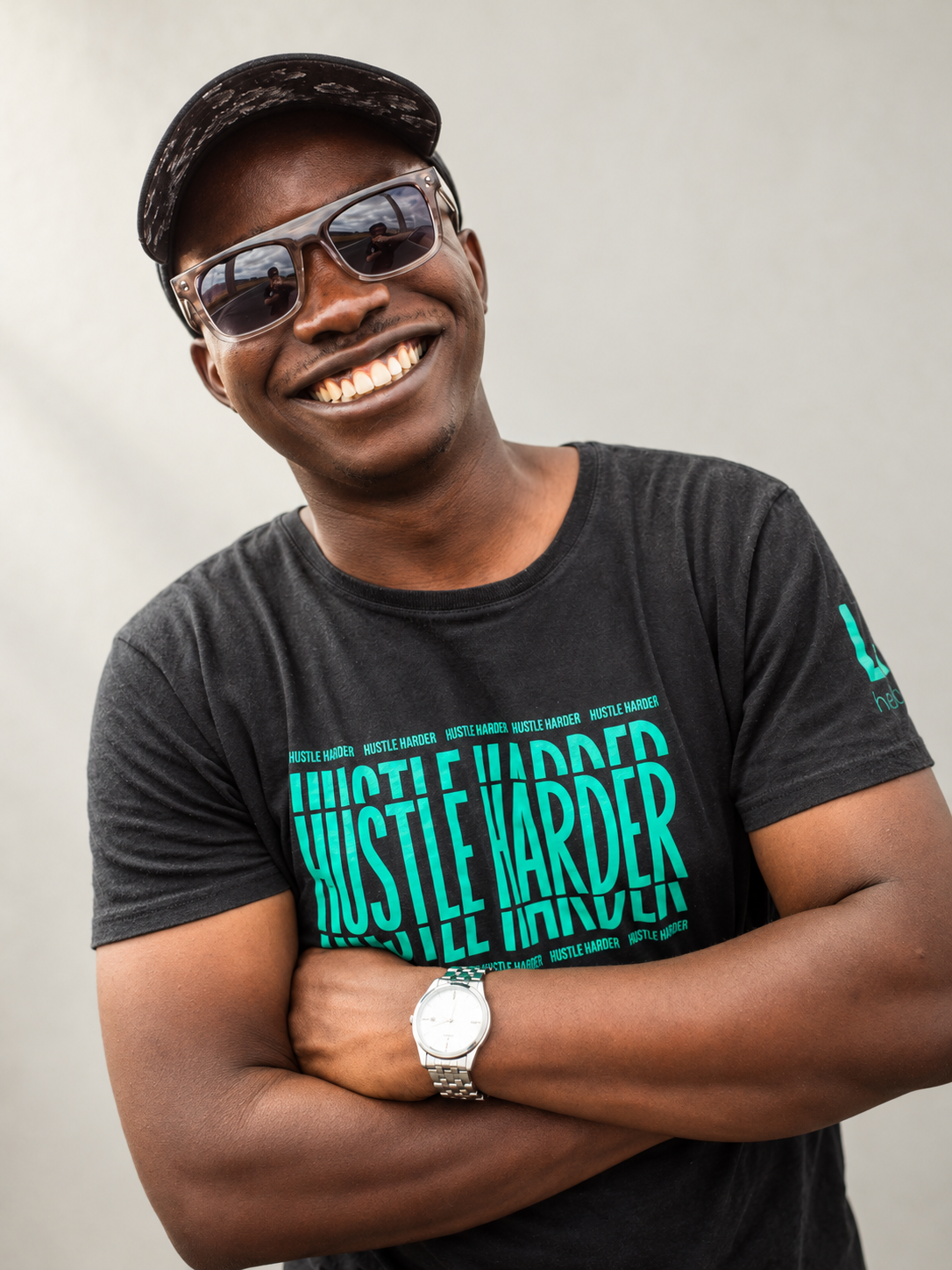 Paul Kidero wearing sunglasses, a black t-shirt with the words HUSTLE HARDER, a white wristwatch, and a black cap, standing on a rural road with a cloudy sky in the background.