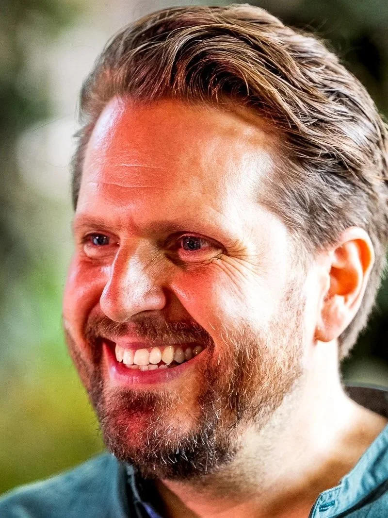 Close-up of Mikkel Becker-Aakervik wearing a blue shirt, outdoors with blurred greenery background.