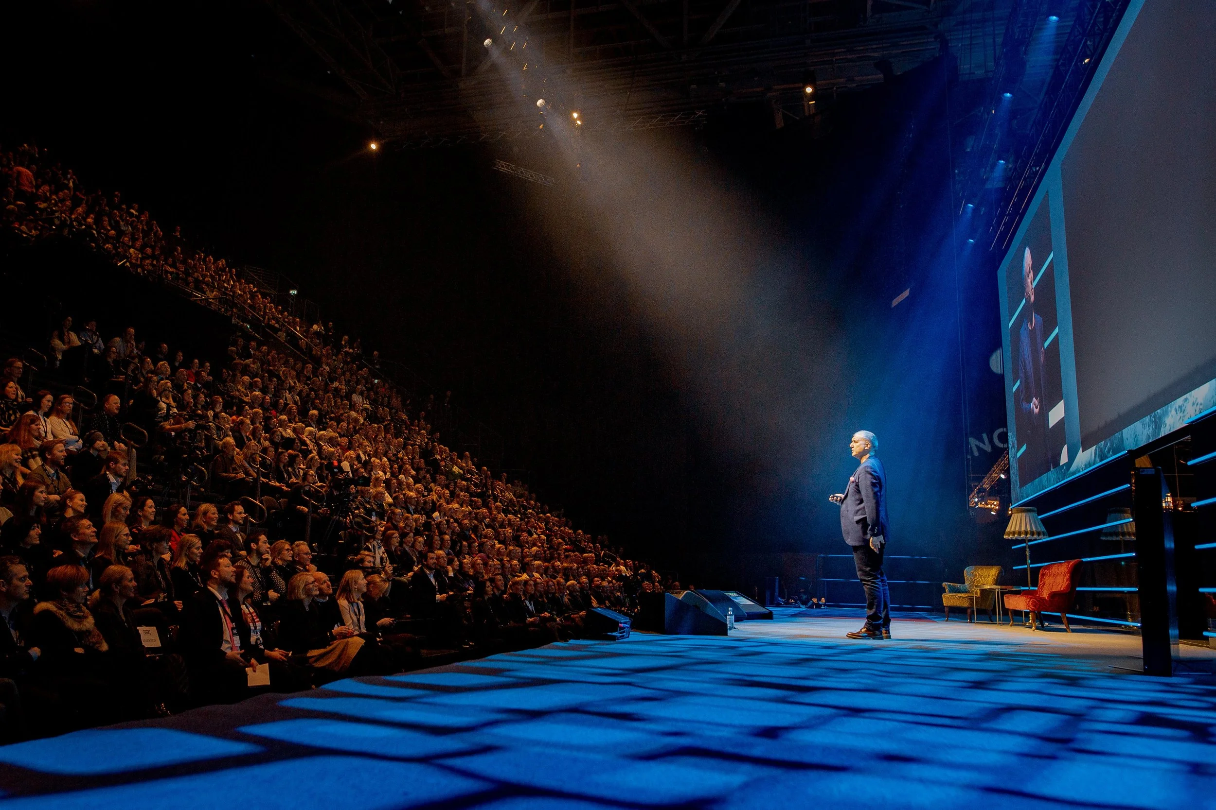 A speaker on stage at a the SHE conference, addressing a large audience in a darkened auditorium. The stage is illuminated with blue lighting, and there are chairs and a lamp on stage. A large screen behind the speaker displays his image.