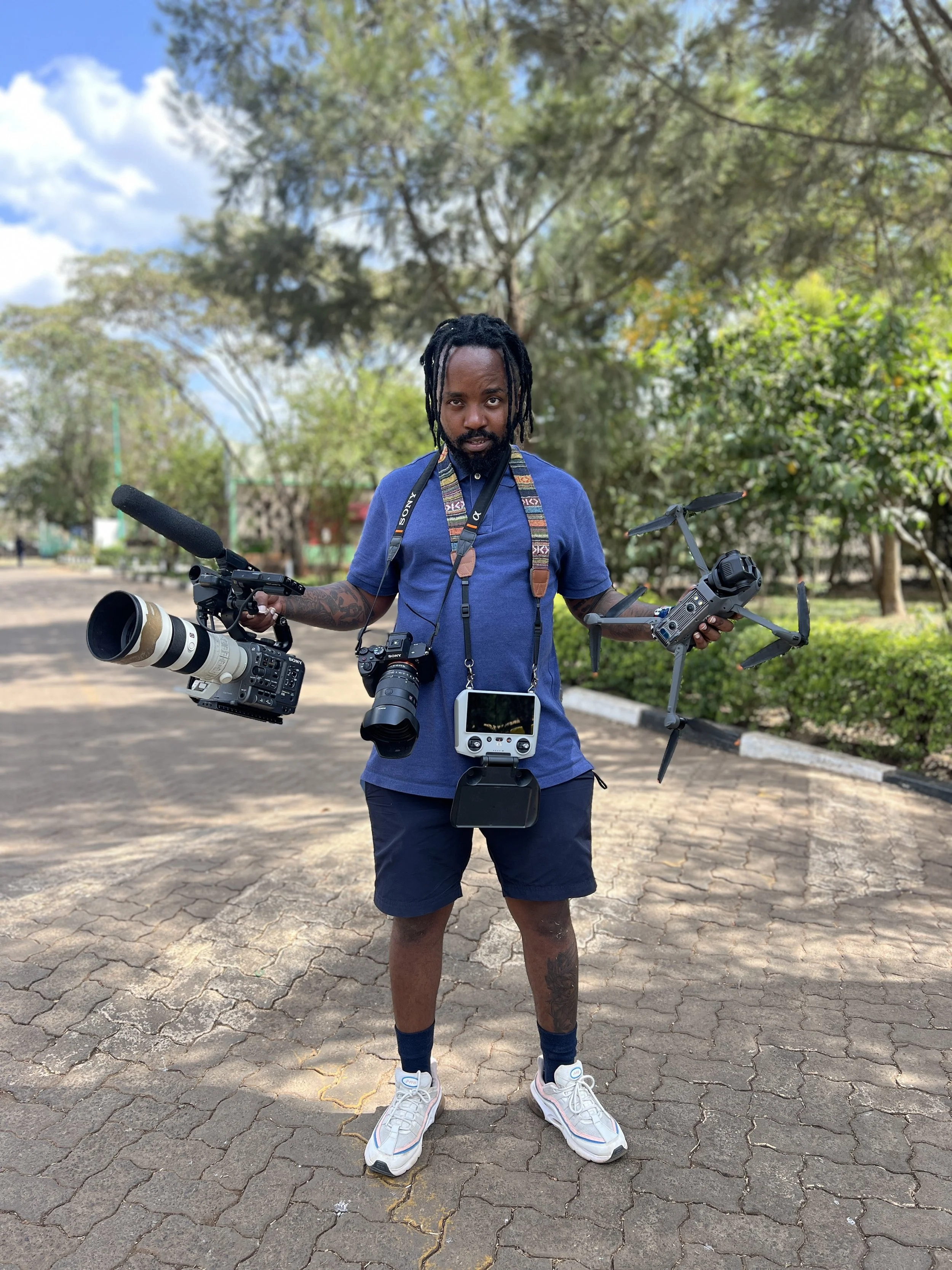 A man standing outdoors on a paved pathway, holding a professional camera with a large telephoto lens, a drone, and a remote control. He is dressed in a blue t-shirt, navy shorts, and white sneakers, with trees and a partly cloudy sky in the background.