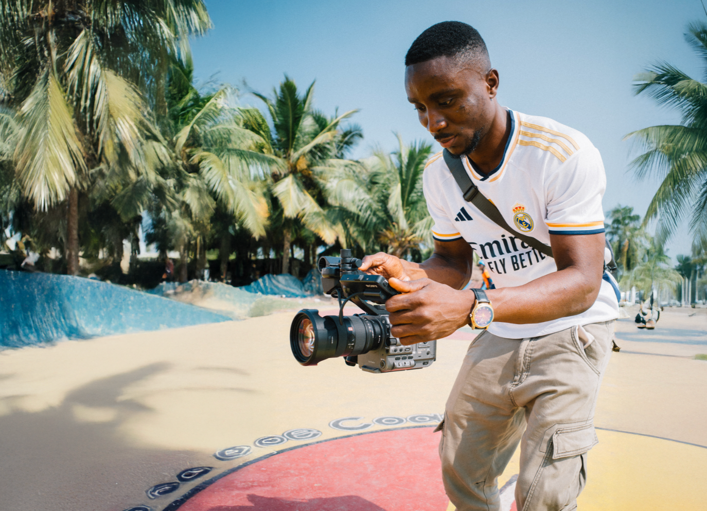 Thought Leader Ghana Lead  member wearing a white Real Madrid soccer jersey and beige cargo shorts operating a professional video camera outdoors near palm trees on a sunny day.