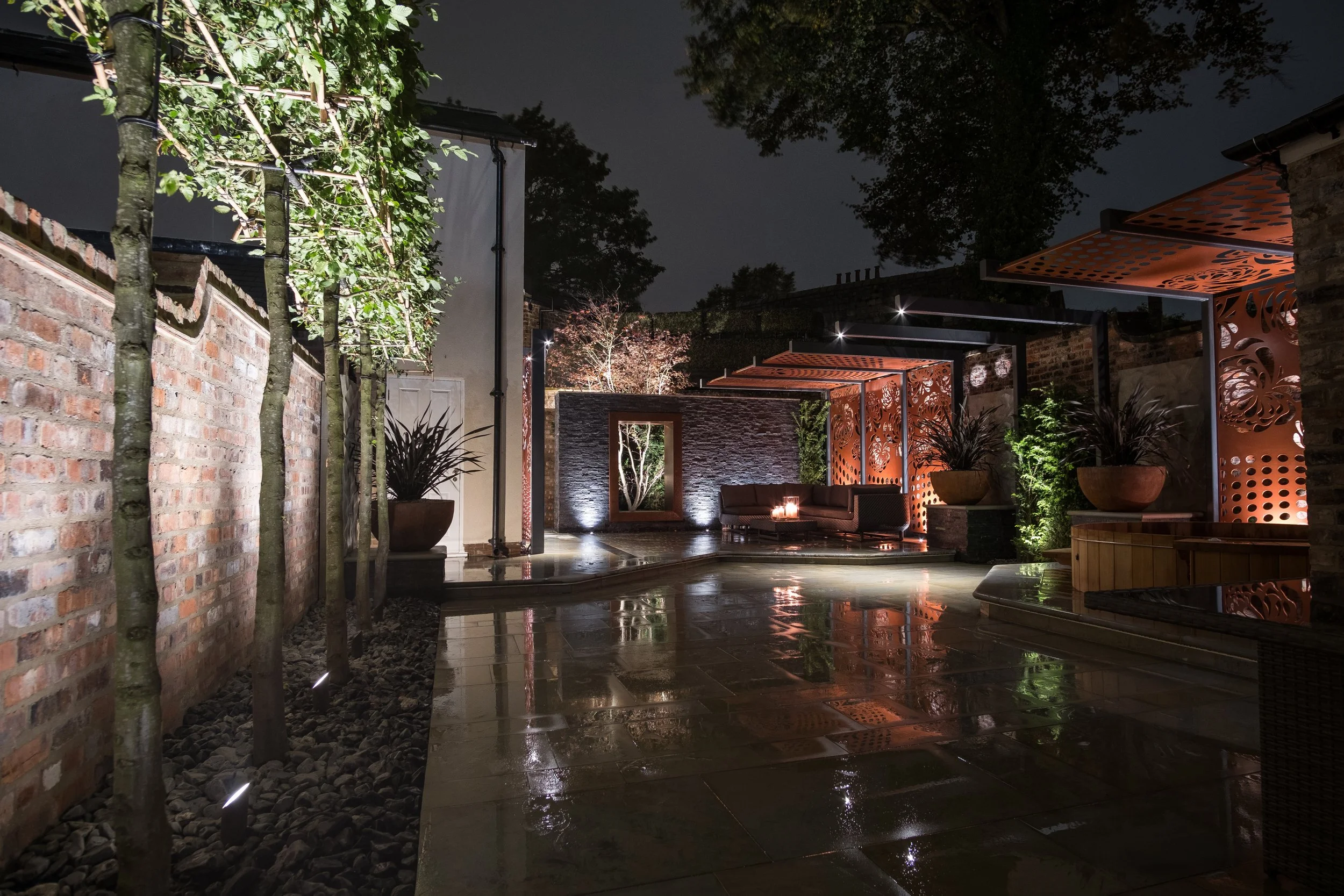 A modern outdoor patio at night with wet stone flooring, orange decorative panels, potted plants, a seating area with a fire pit, trees, and ambient lighting.