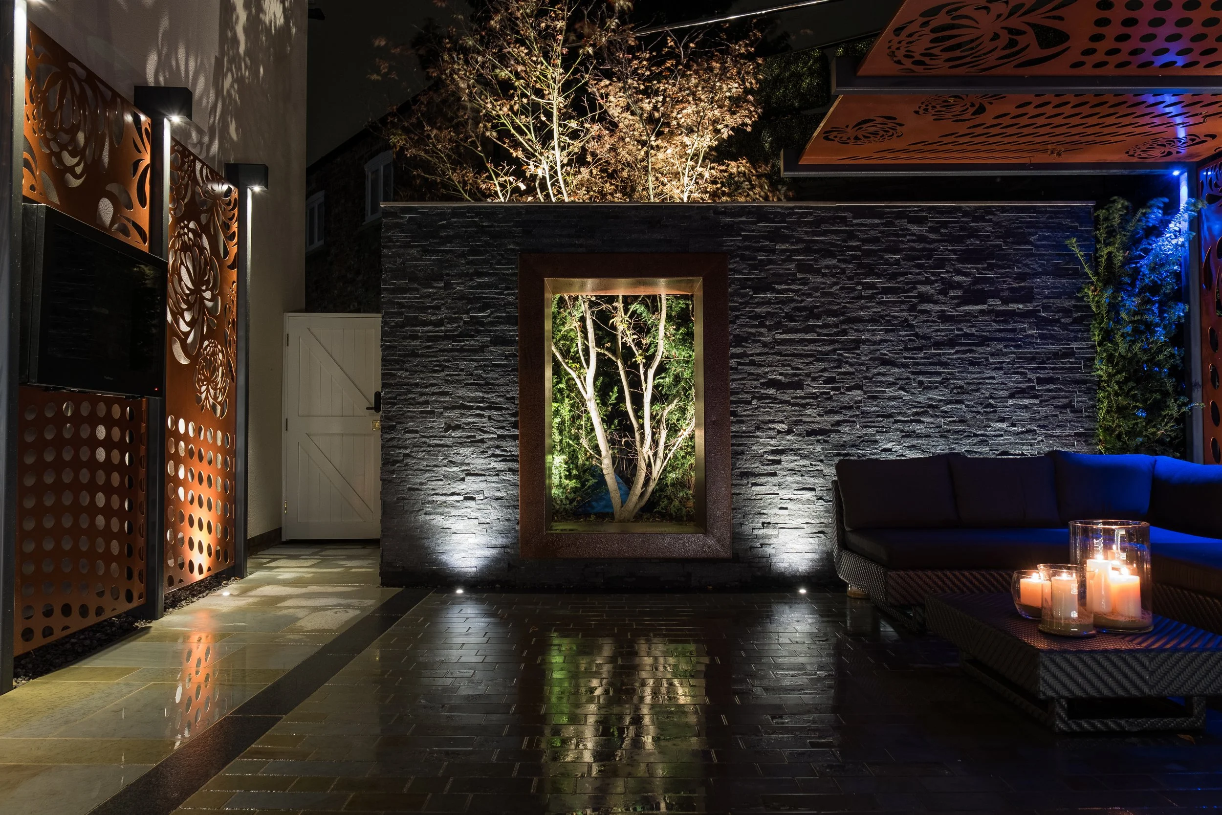 A modern outdoor patio area at night with a black textured brick wall, a window with a tree inside lit from behind, candles on a wicker table, and decorative laser-cut orange panels on the ceiling and walls.