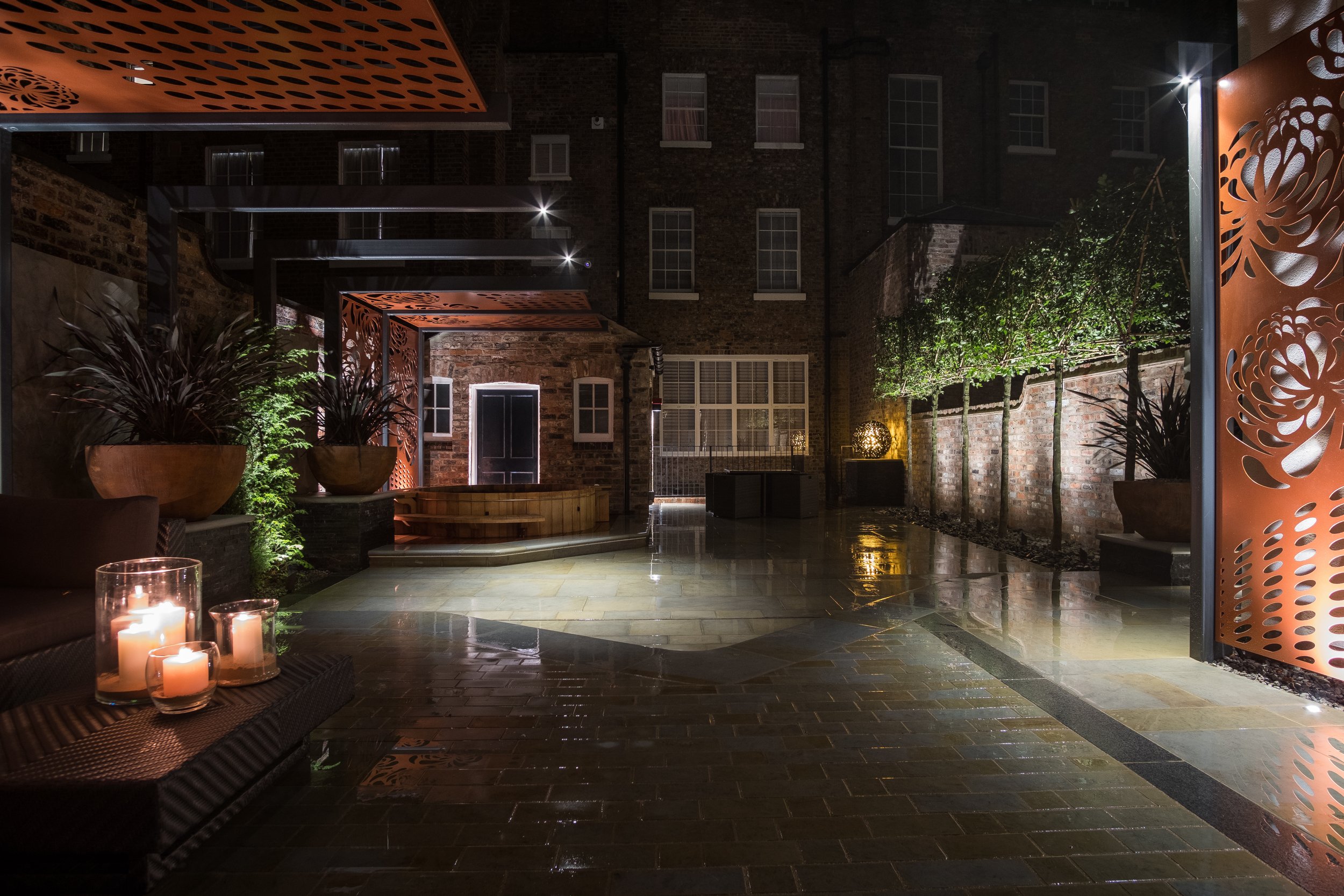 An outdoor patio at night with lit candles on a bench, potted plants, decorative orange metal screens, and a brick wall with windows. The ground is wet and reflects lights.