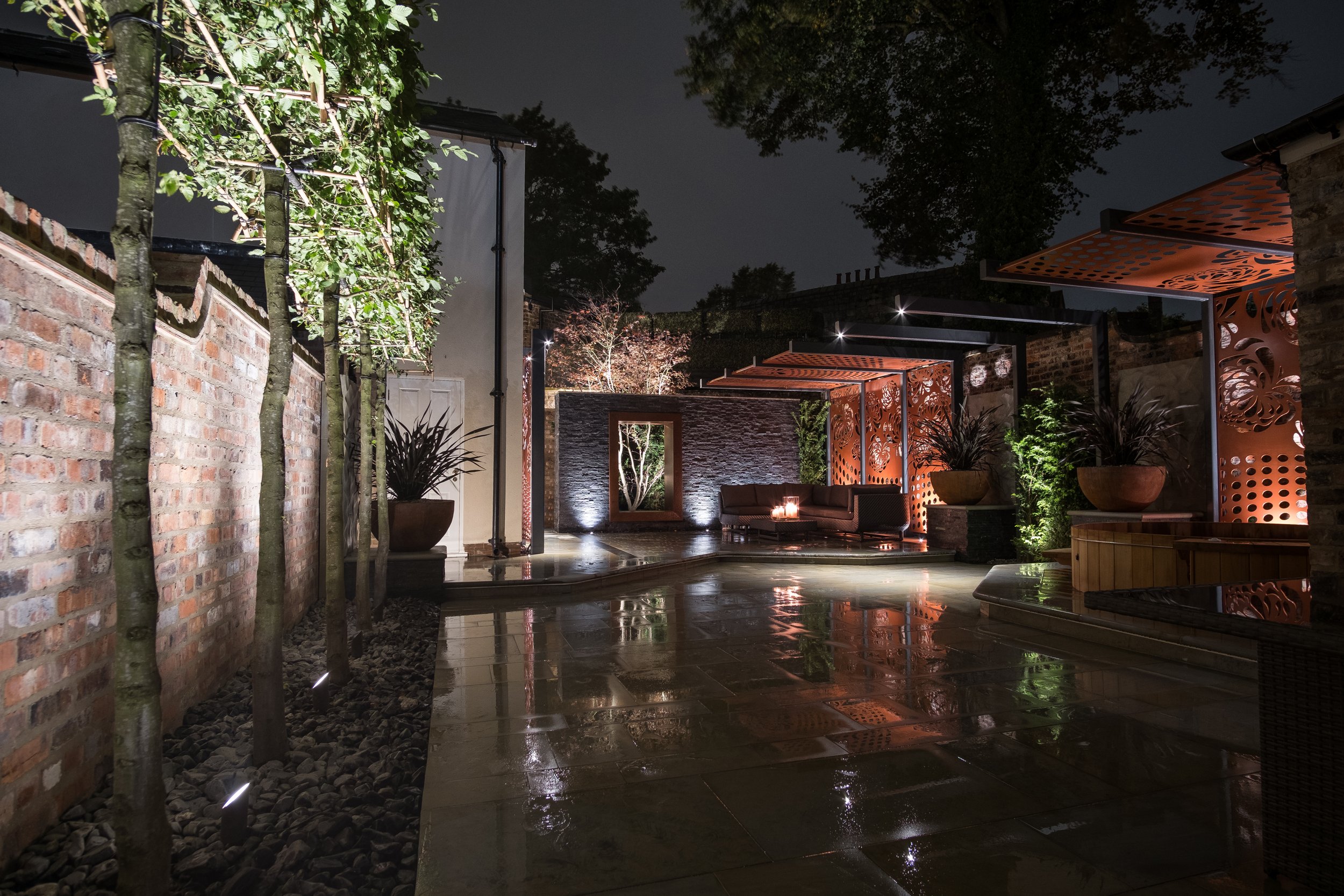 Night view of a modern outdoor patio with wet stone flooring, brick and stone walls, illuminated trees, potted plants, and a seating area with a fire pit, decorated with orange lattice panels.