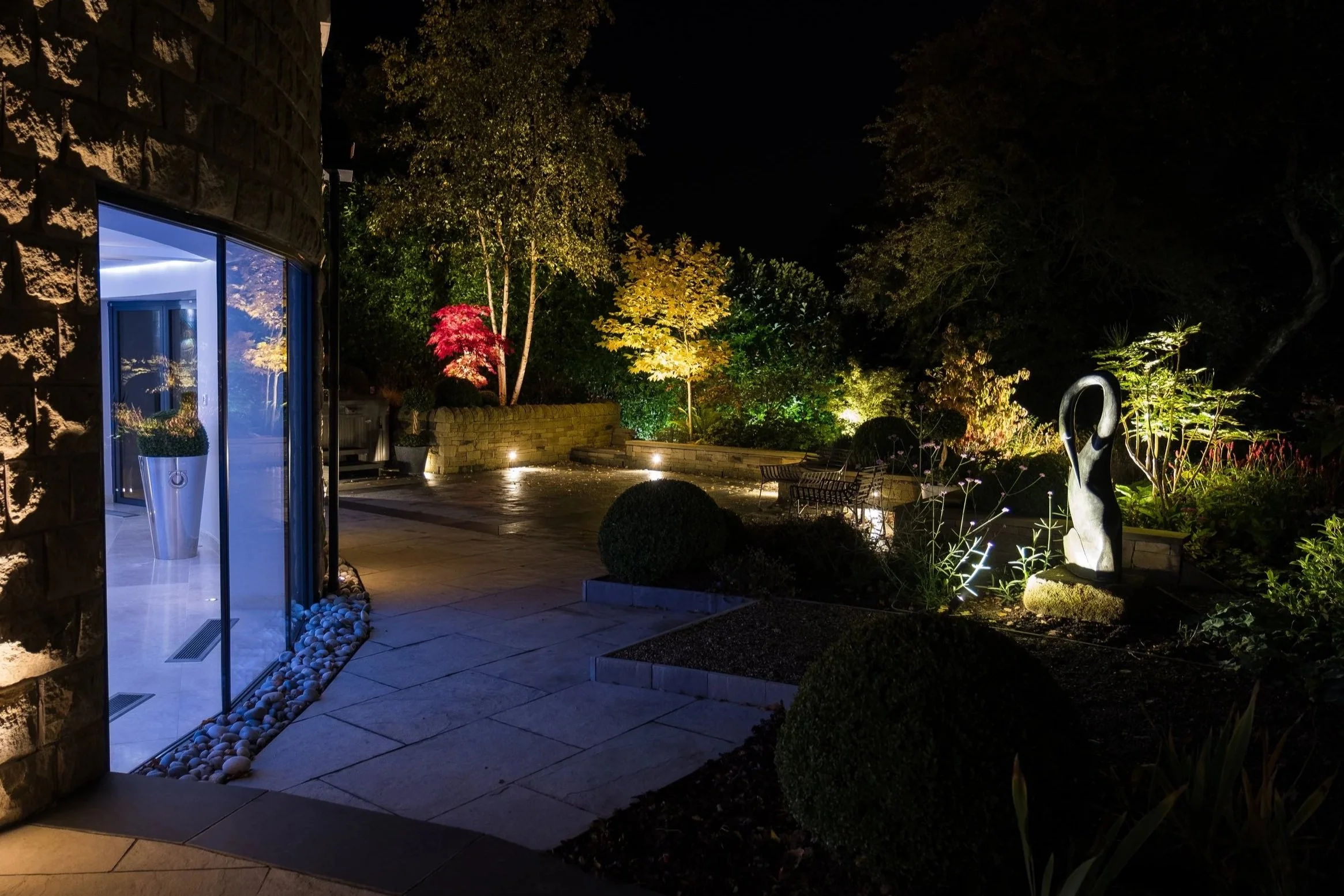 Nighttime view of a modern backyard with illuminated trees, garden sculptures, and outdoor seating, seen from a patio with a glass door.