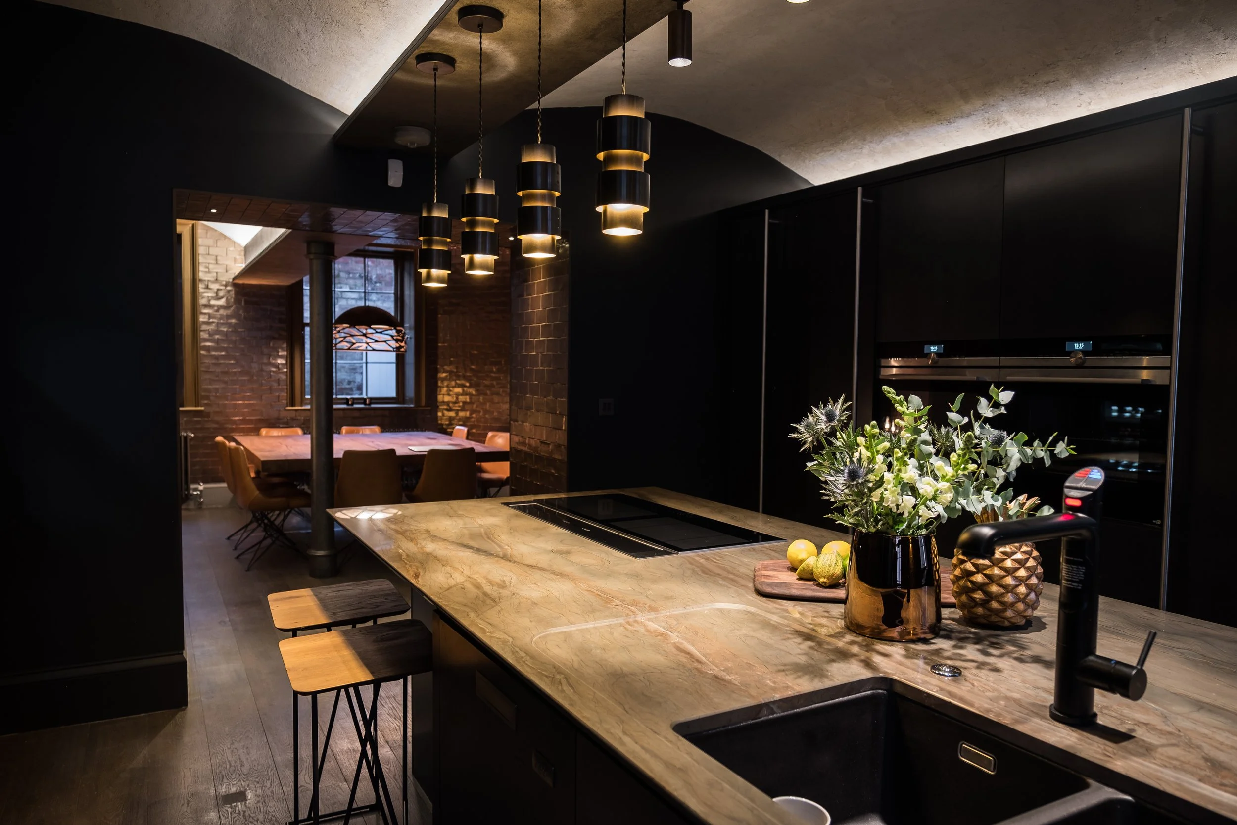 Modern kitchen with marble countertop, black cabinets, and a vase with flowers. In the background, there is a dining area with a large wooden table and chairs, brick walls, and hanging pendant lights.