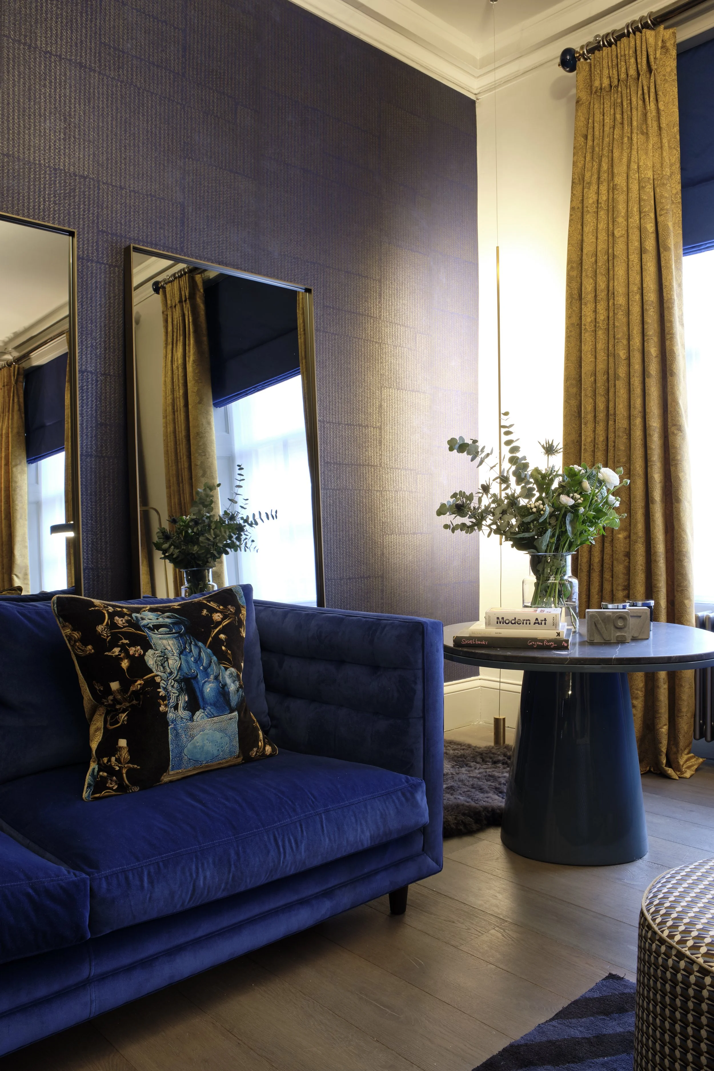 Living room with deep blue velvet sofa, black round side table with a flower arrangement, books, and a camera, large windows with gold curtains, and two tall mirrors on a textured blue wall.