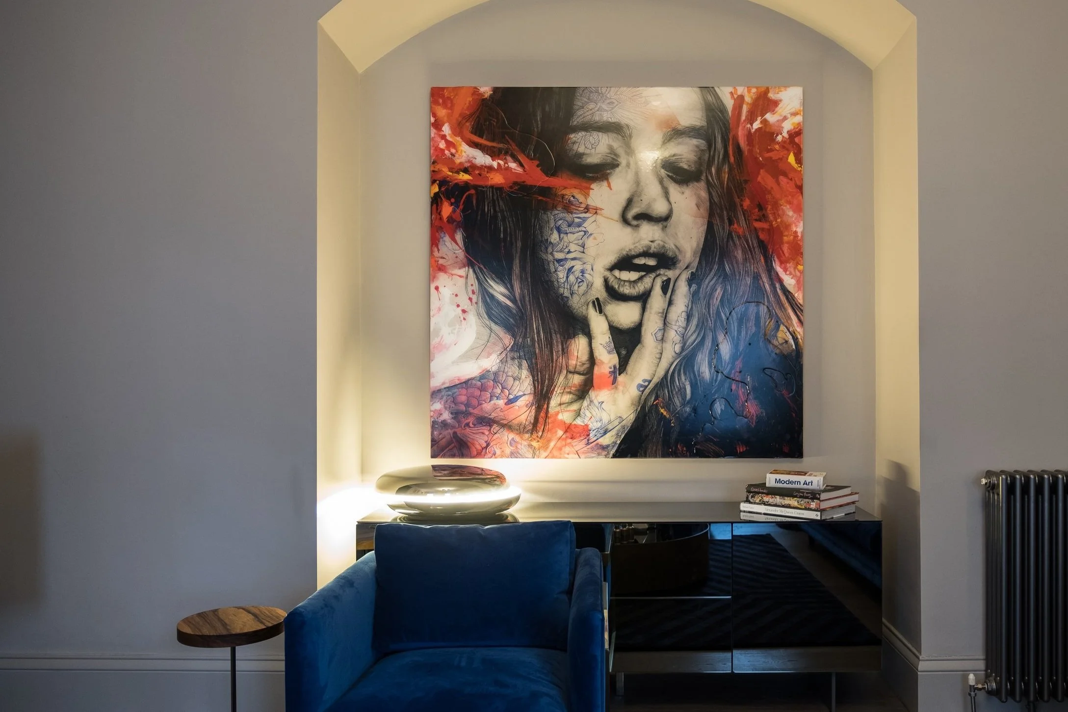 A living room with a large contemporary artwork of a woman's face, partially in black and white, with colorful abstract paint splashes in red, orange, yellow, and blue. The artwork hangs on a wall within a recessed, curved wall niche. Below the artwork, there is a black sideboard with a stack of books, including one titled 'Modern Art,' and a white, layered decorative object. To the left, a blue velvet armchair is partially visible, and a small round wooden side table is next to it. On the right side of the image, a black radiator is seen.