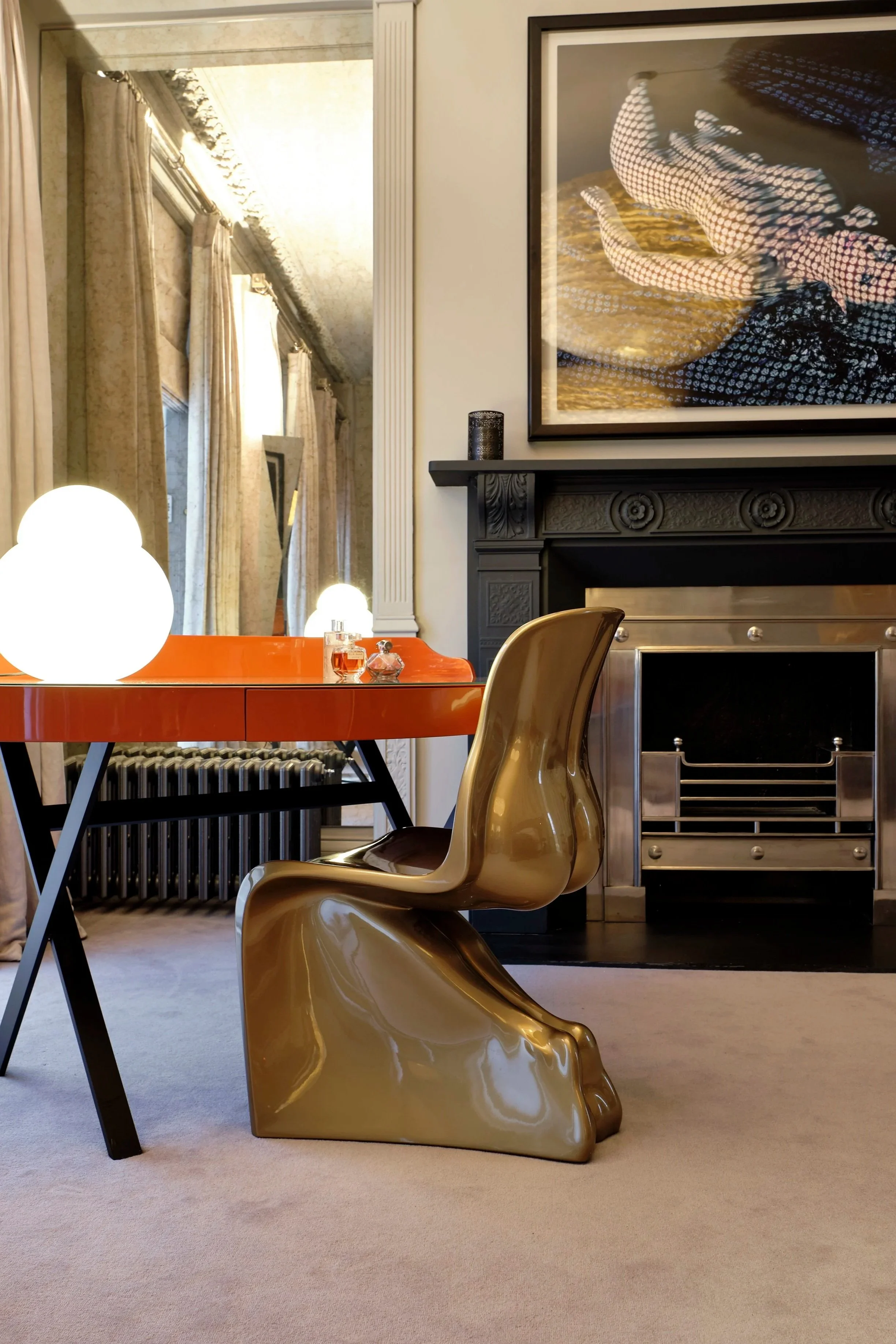 Interior of a room with a black fireplace, framed artwork, orange table, gold modern chair, and two glowing white table lamps with curtains and windows in the background.