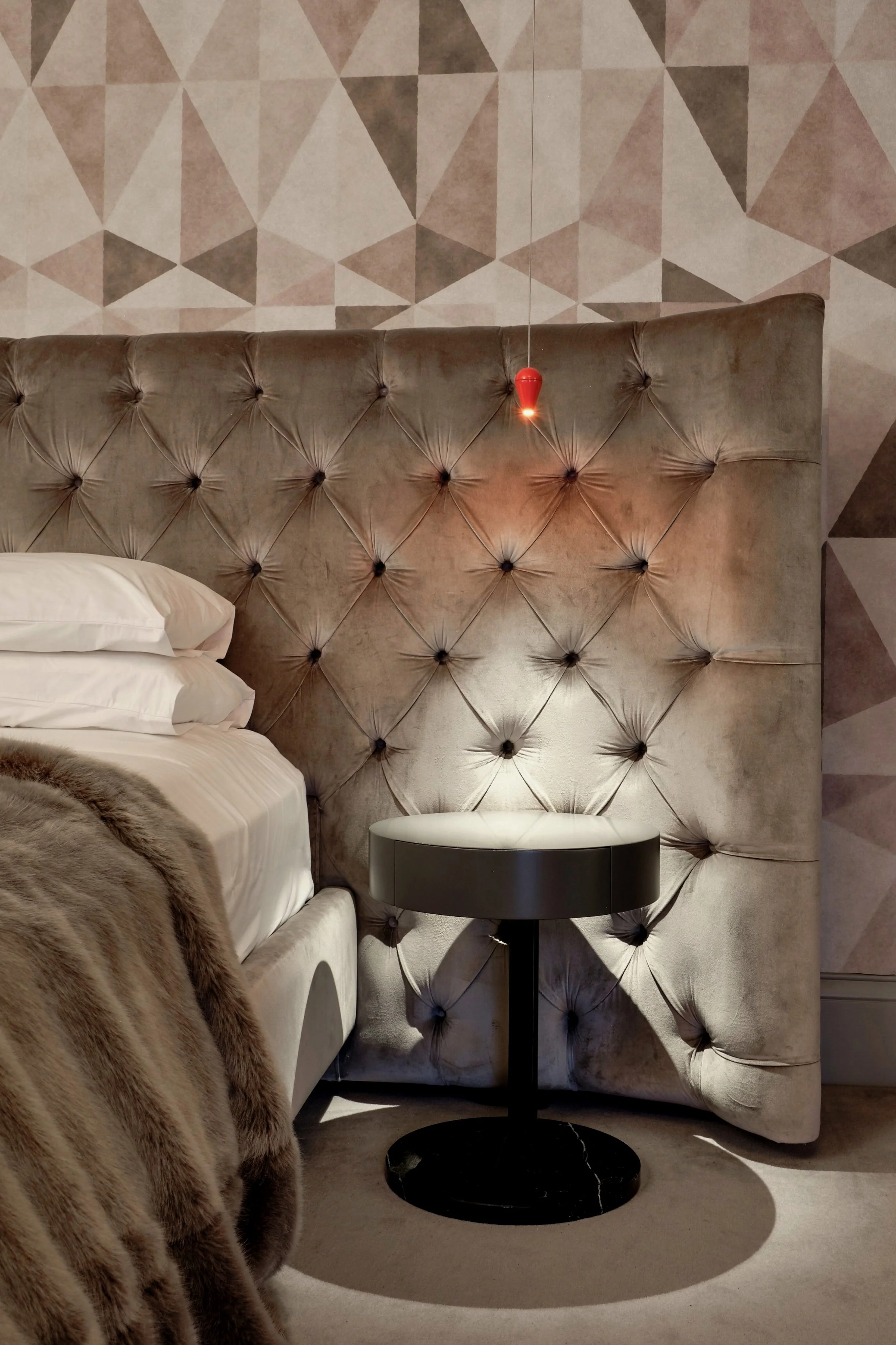 A bedroom with a large tufted velvet headboard, a nightstand, and a partial view of a fur blanket on the bed. There is a geometric patterned wallpaper on the wall and a small red pendant light hanging above the headboard.