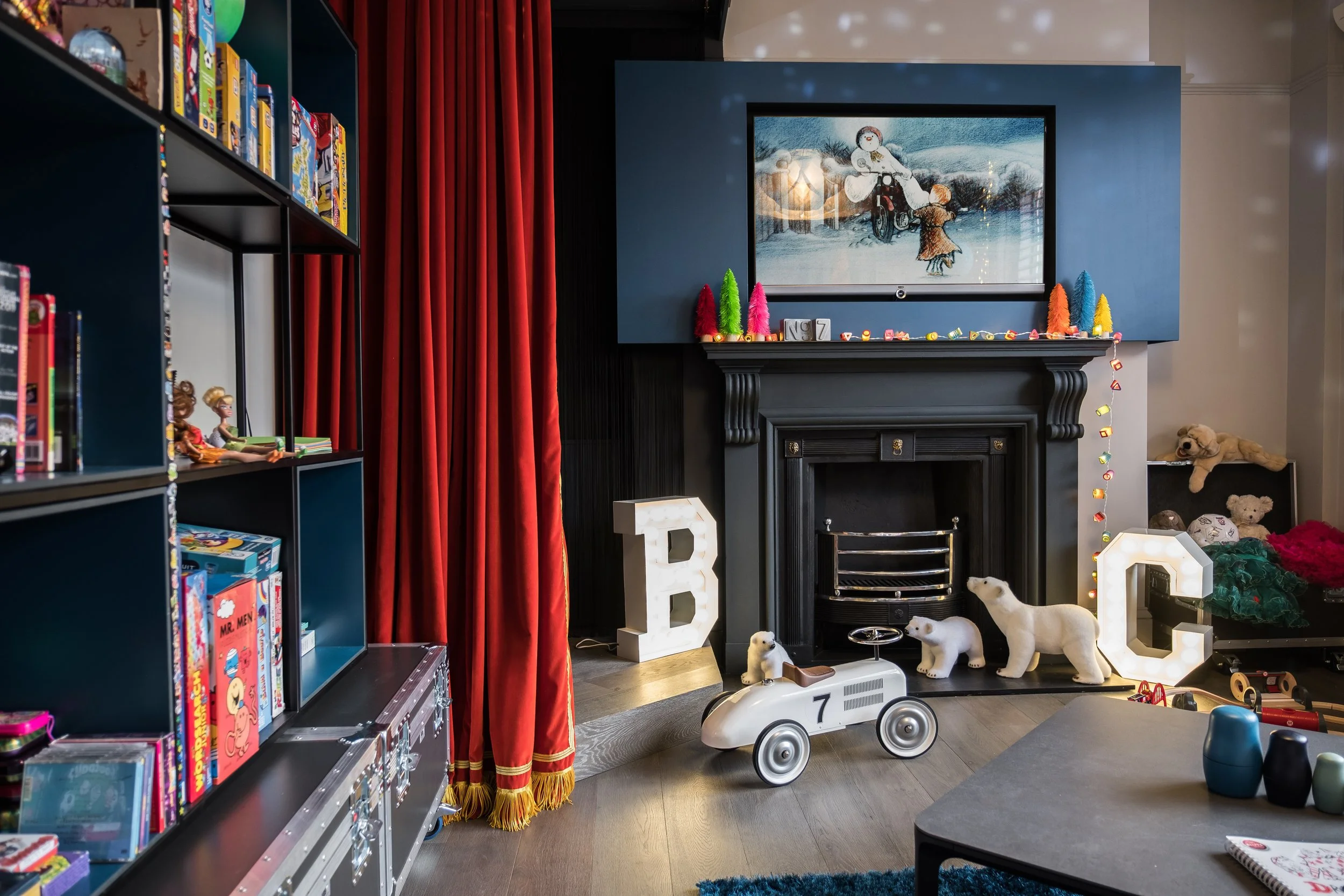 Living room with a black fireplace, decorated with colorful lights and paper trees, with lit-up letters 'B' and 'C' on either side. A television shows a snowman scene. Toys, including plush polar bears and a toy car, are scattered around. Red curtains cover a window on the left.