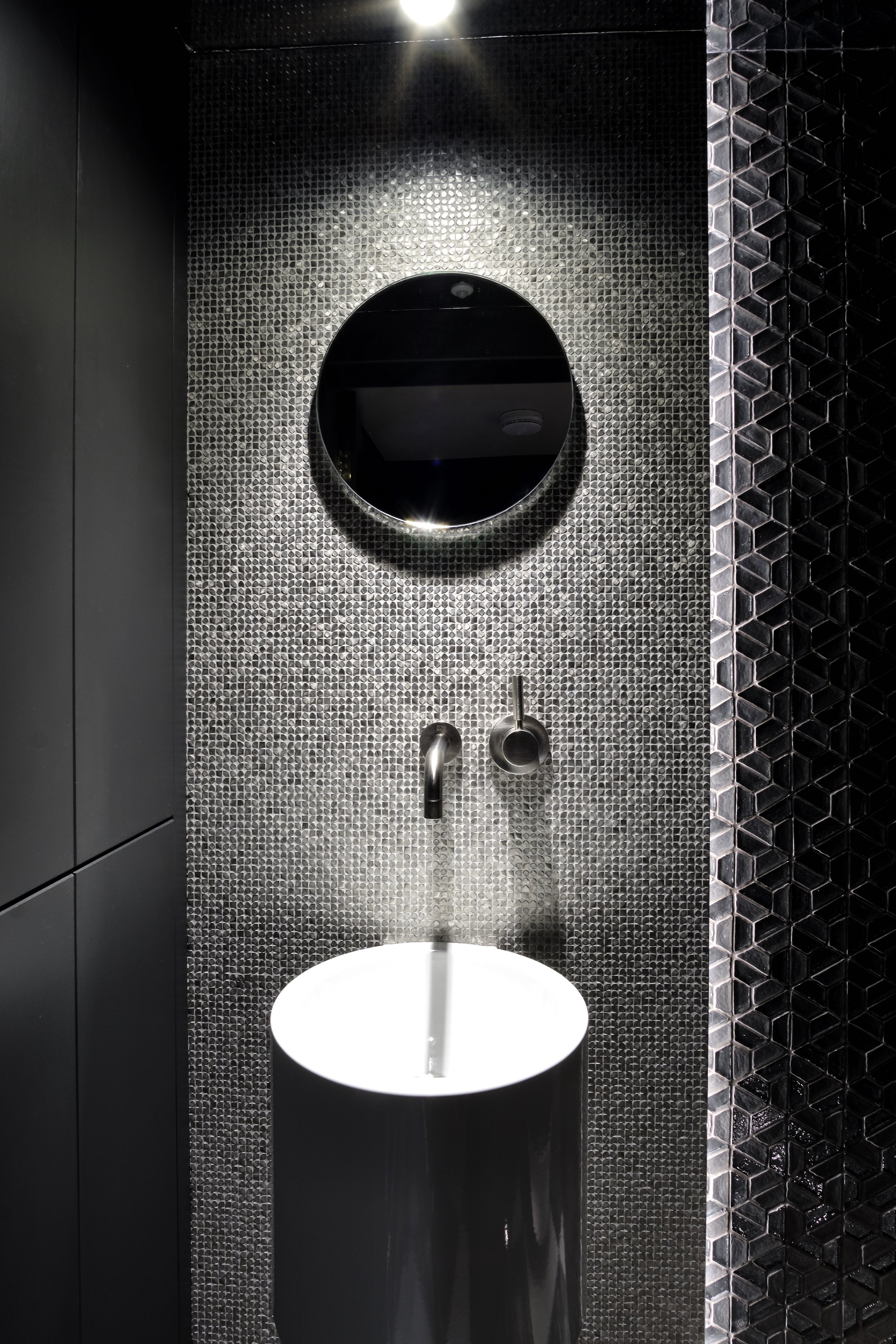 Modern bathroom with black and metallic decor, a round mirror, and a cylindrical white sink with a wall-mounted faucet.