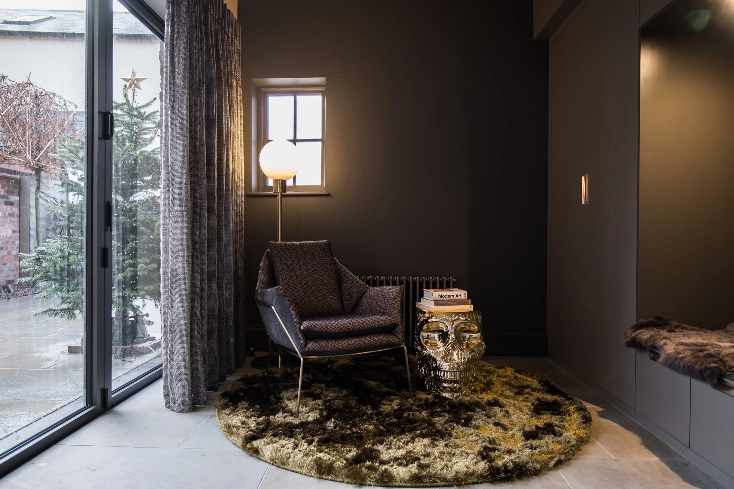 A cozy corner in a modern living room with dark walls, a chair, a floor lamp, a small side table with books and a decorative skull, a circular rug, and sliding glass doors leading outside.