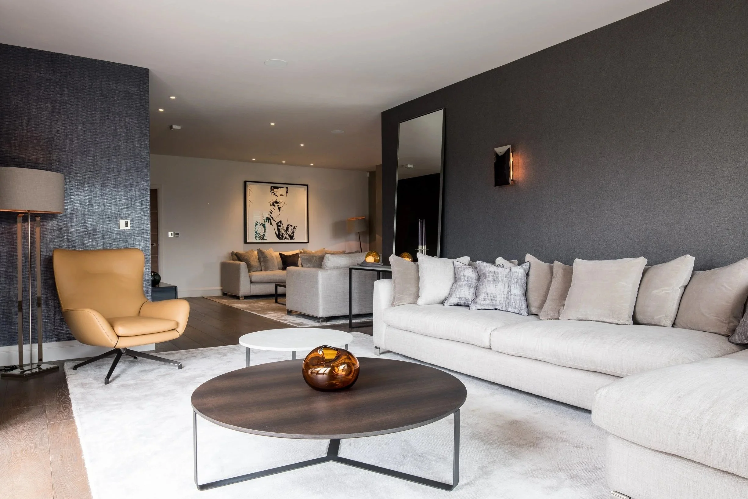 Modern living room with beige couches, a tan armchair, dark wood coffee tables, and a white area rug. There is a large framed black-and-white portrait on the wall and a black textured accent wall.