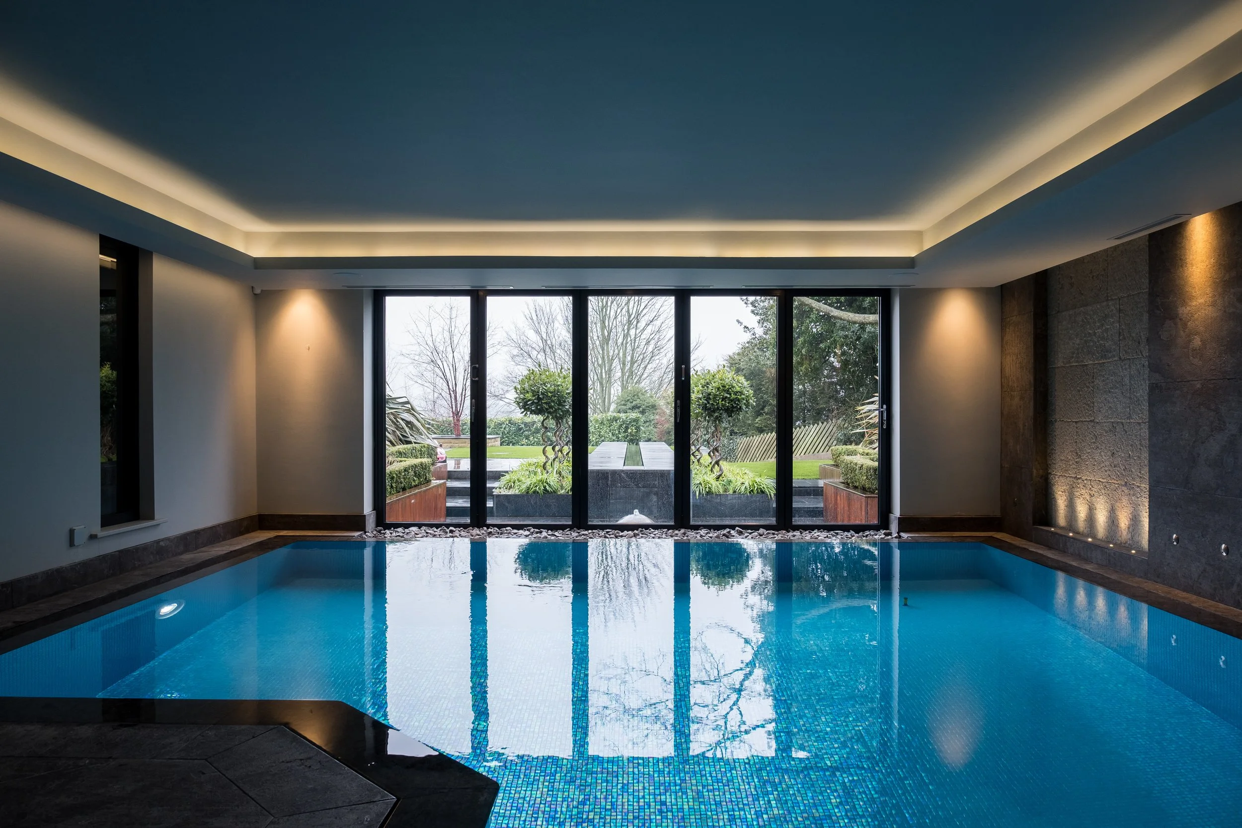 Indoor swimming pool with large glass doors showing a garden outside, modern ceiling lighting, and stone accents on the wall.