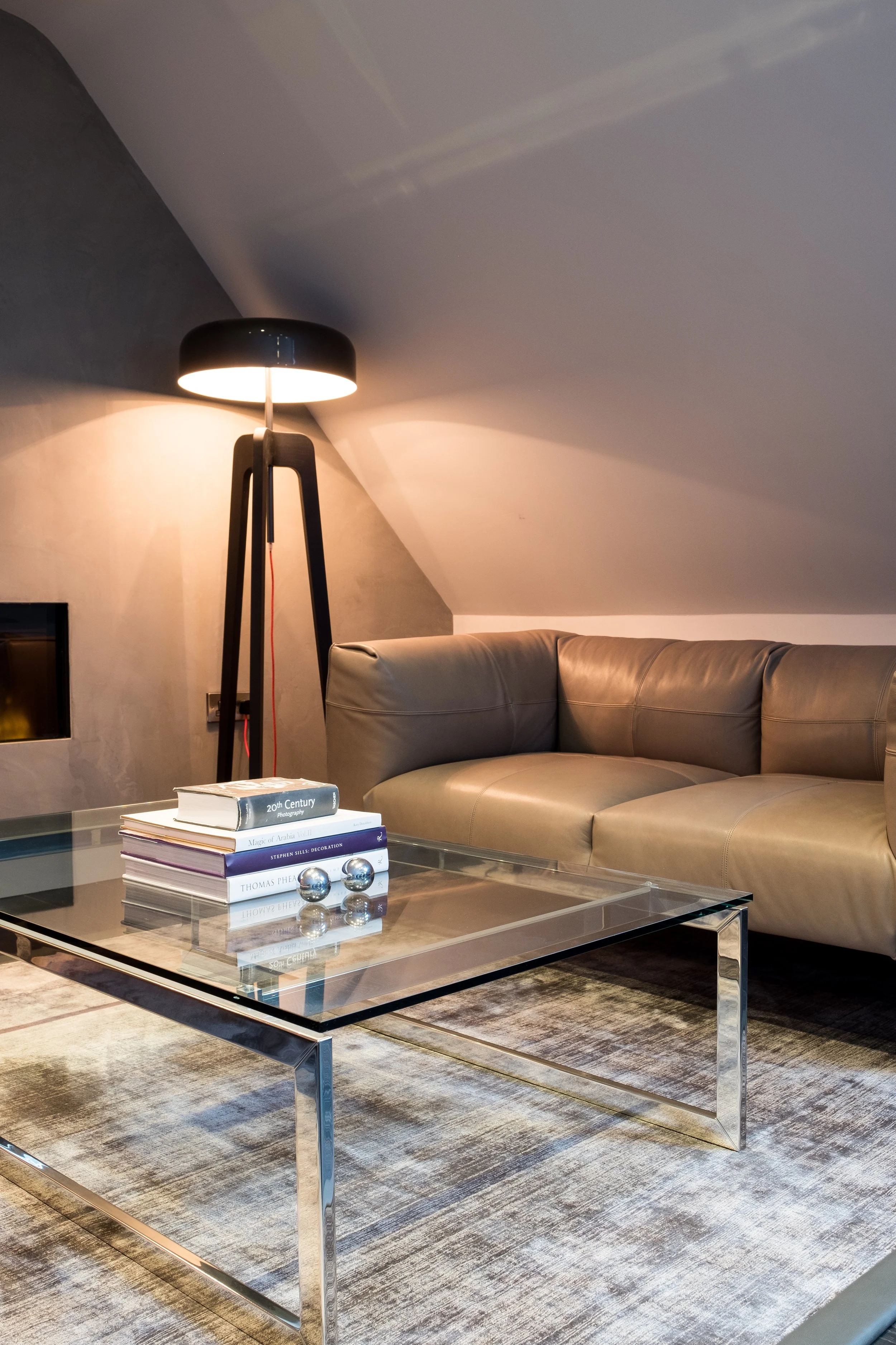 Modern living room with a black floor lamp, beige leather sectional sofa, glass coffee table, books, and decorative metal spheres.