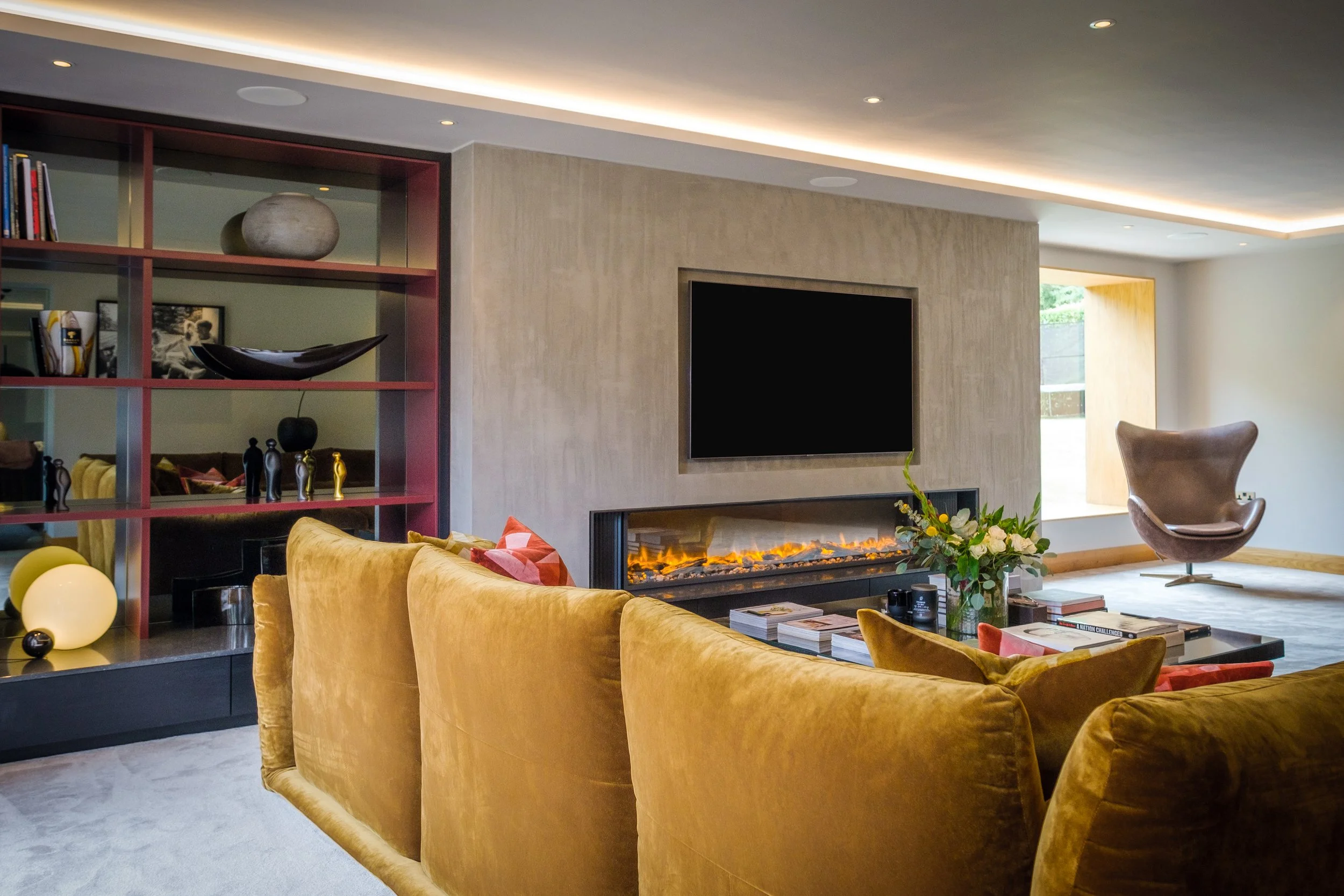 Modern living room with a mustard velvet sofa facing a wall-mounted TV and electric fireplace, decorative shelves on the left with sculptures and books, and a rocking chair near a window.