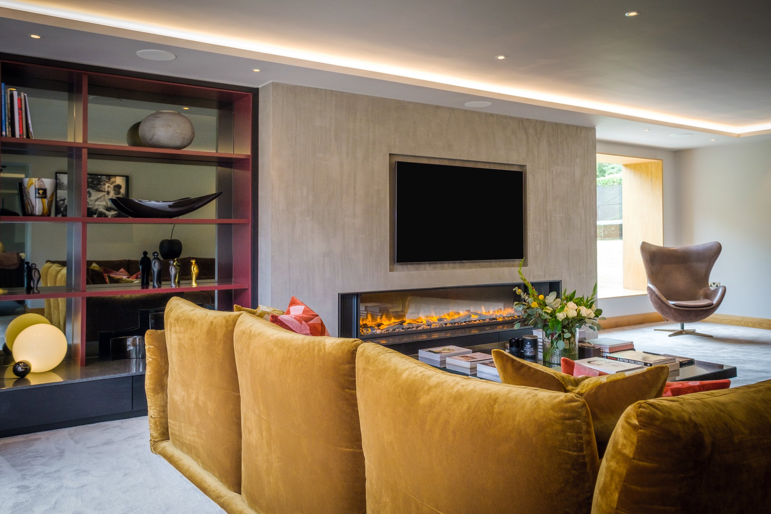 A modern living room with a yellow velvet sectional sofa, a flat-screen TV above a fireplace, a purple lounge chair near a window, a black and red shelf with decorative items, and a coffee table with books and a flower arrangement.