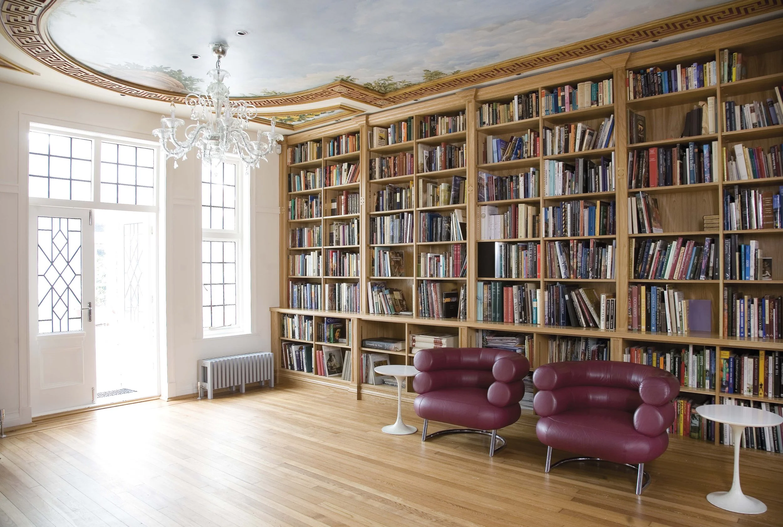 A cozy library room with large windows, a white door, a chandelier, tall wooden bookshelves filled with books, two burgundy chairs, and small white side tables.