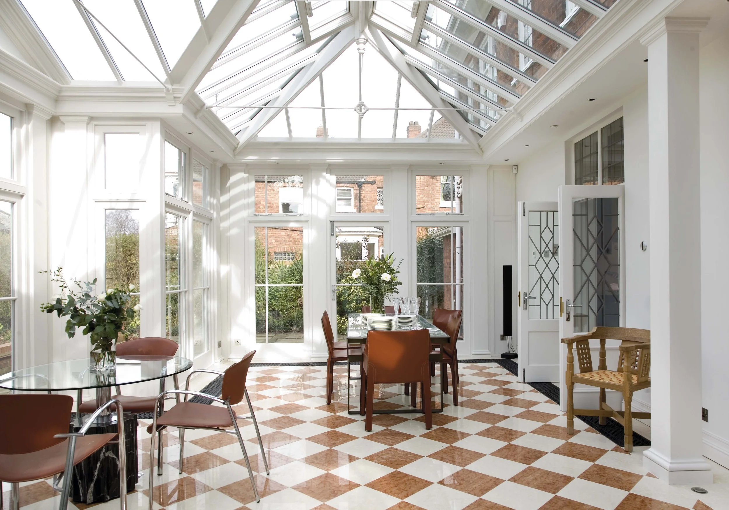 Bright sunroom with a glass ceiling, white walls, and a checkered tile floor. Contains a glass table with chairs, a wooden dining table with chairs, and decorative plants.