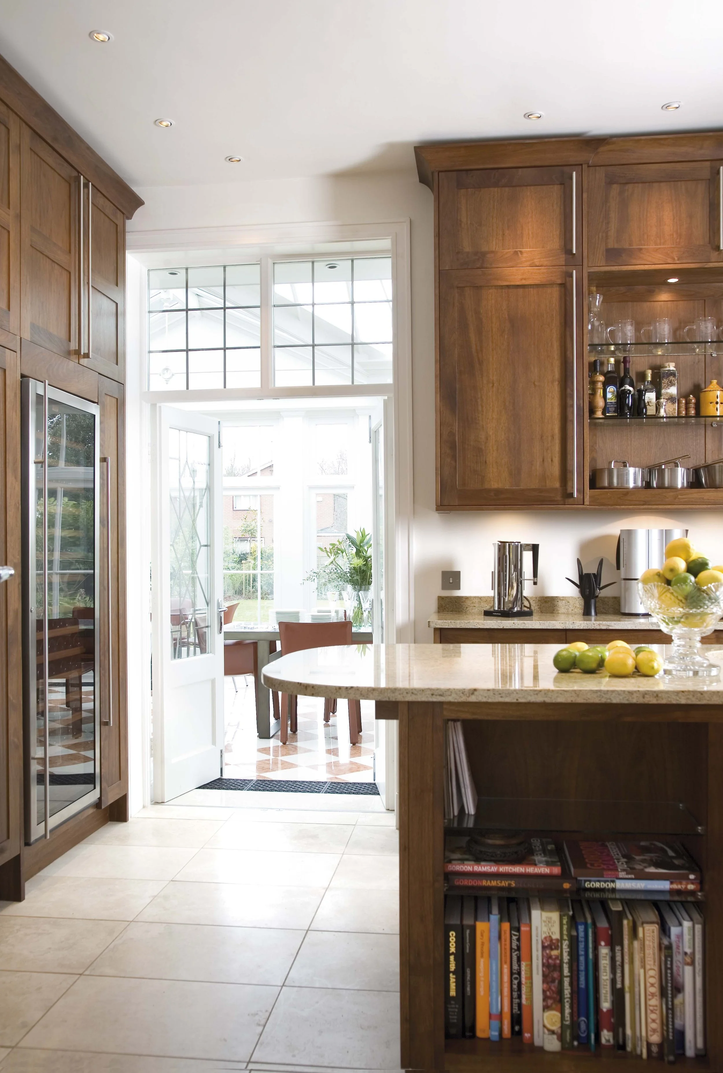 Kitchen with wooden cabinets, a curved granite countertop, a bowl of green and yellow lemons, a glass door leading to a sunroom, and open shelving with glassware and bottles.
