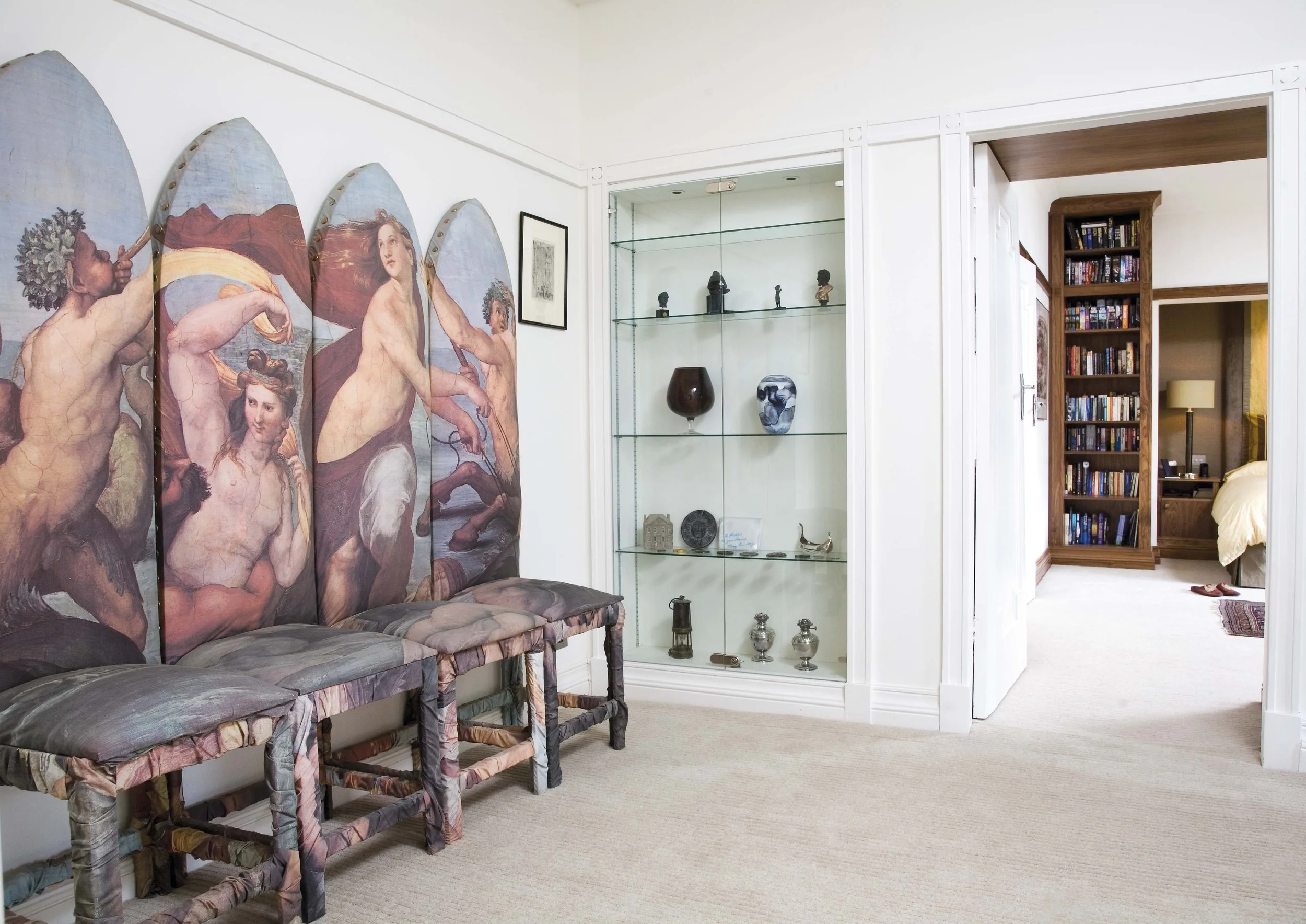 A decorated room with a painted folding screen depicting mythological characters, three chairs with fabric-covered seats, a glass display cabinet with small sculptures and vases, and an open doorway to a bedroom with a bookshelf and a yellow wall.
