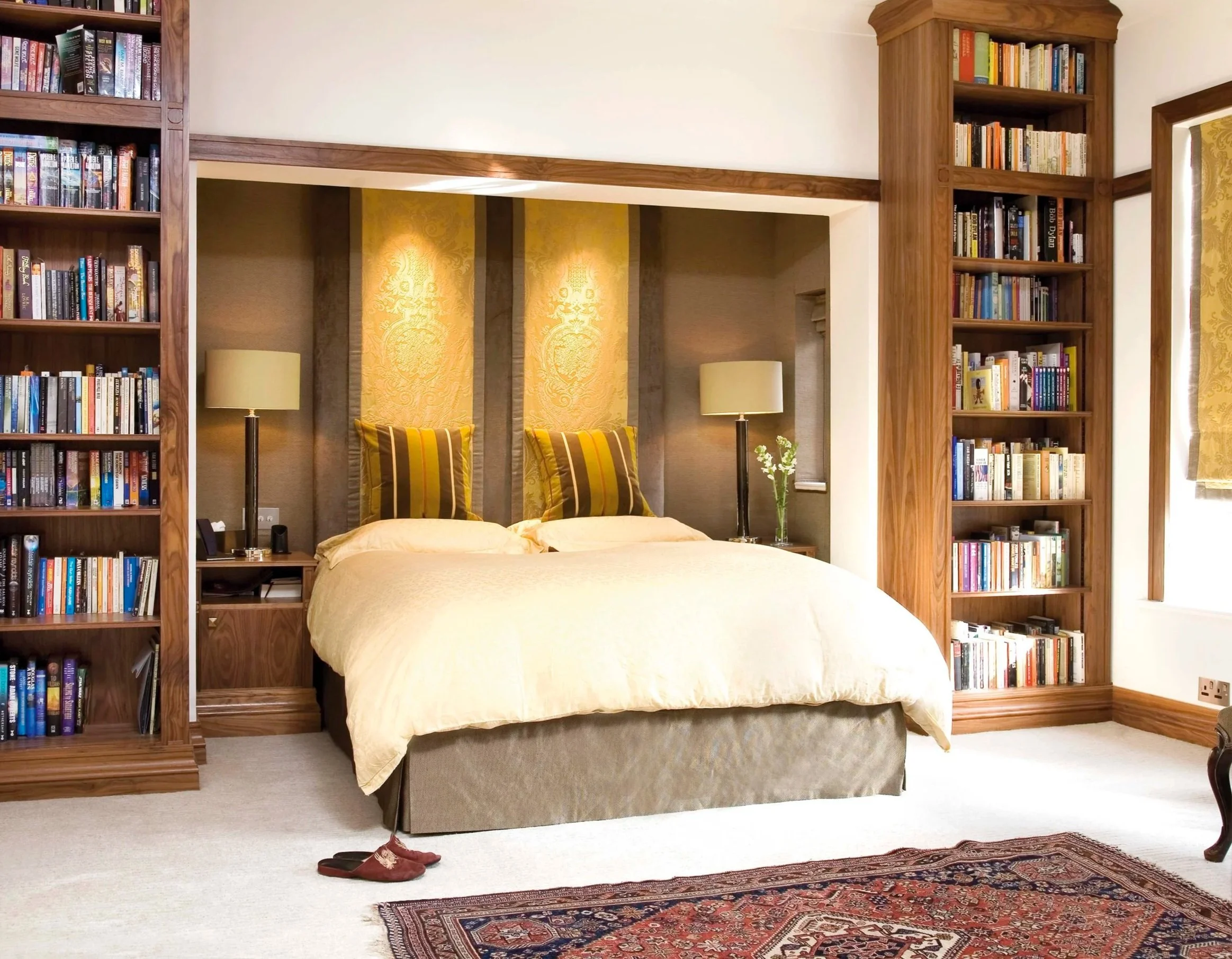 A cozy bedroom with a bed, two nightstands with lamps, built-in wooden bookshelves on either side, a window with curtains, a carpet, and slippers on the floor.