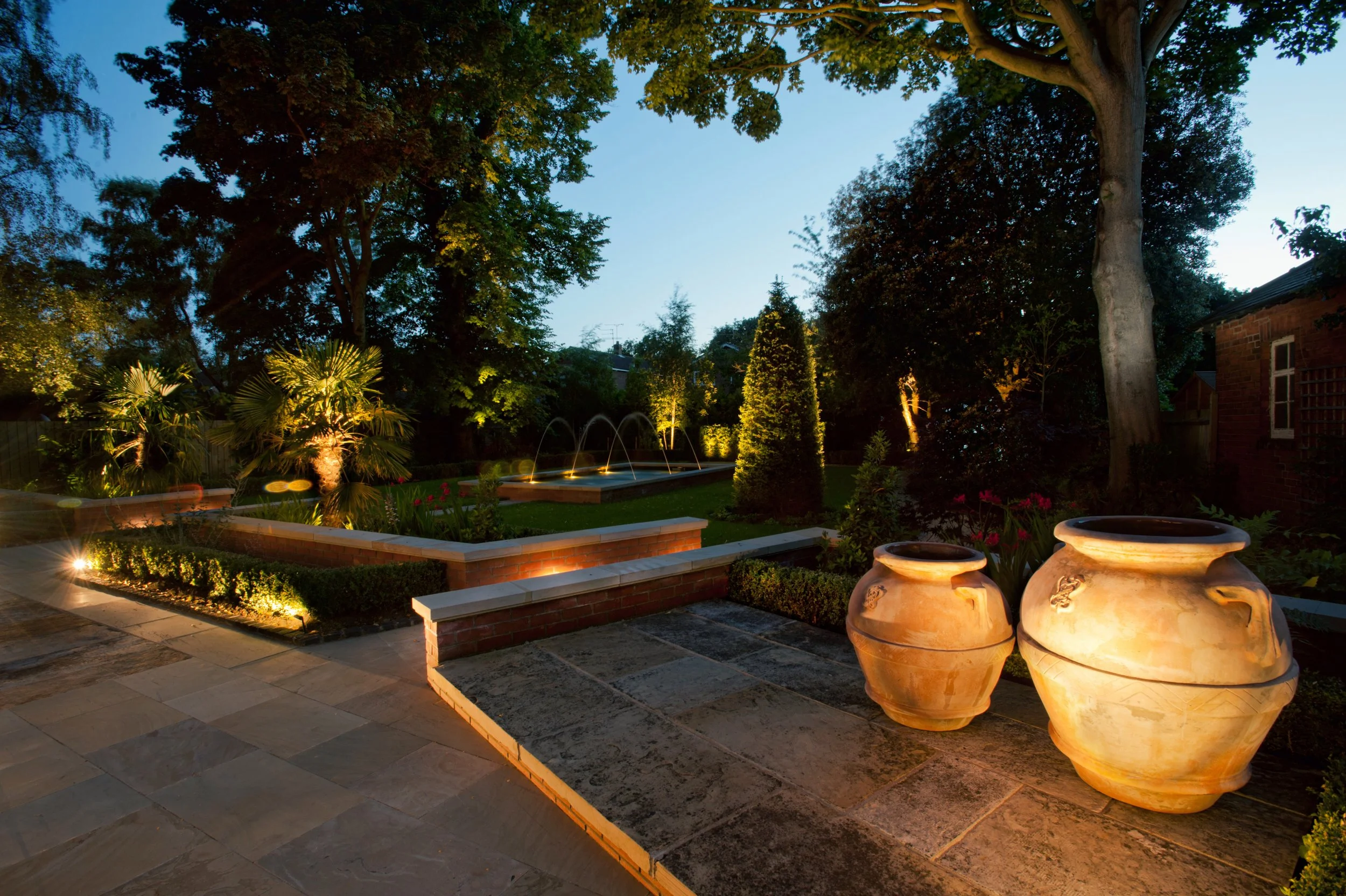 Nighttime view of a landscaped garden with large clay pots, illuminated bushes, a water fountain with arches, tall trees, and a brick building in the background.
