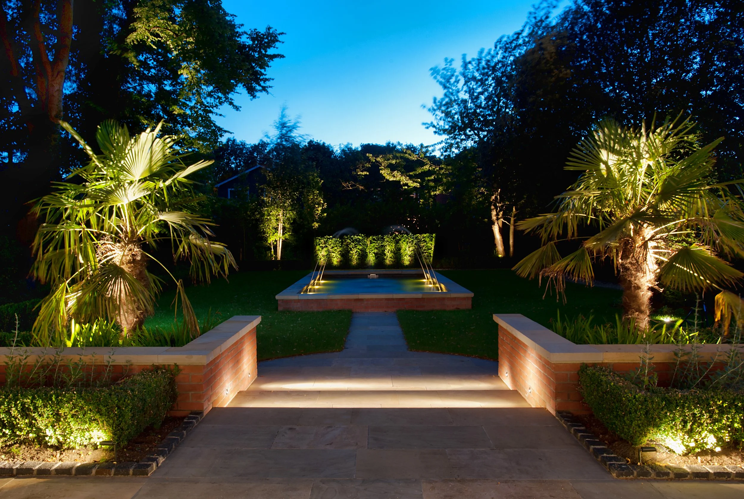 A backyard with a small fountain, surrounded by palm trees, bushy plants, and trees, lit up by landscape lighting at dusk or early evening.