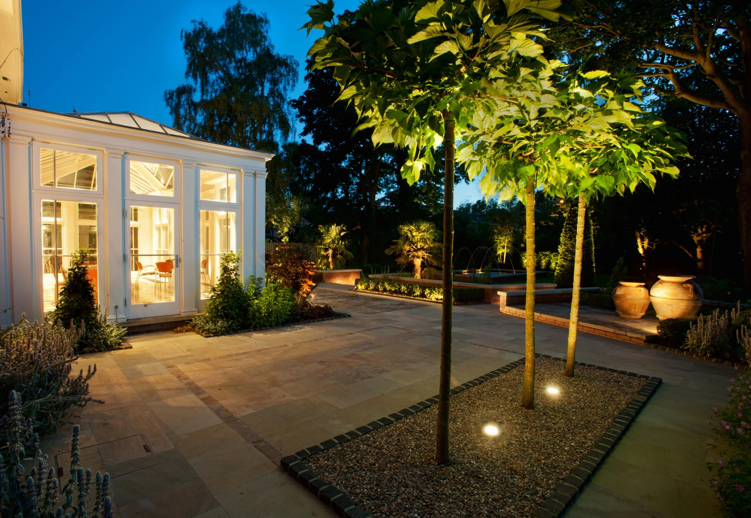 A beautifully lit backyard with trees, potted plants, and a modern greenhouse or sunroom with large windows at nighttime.