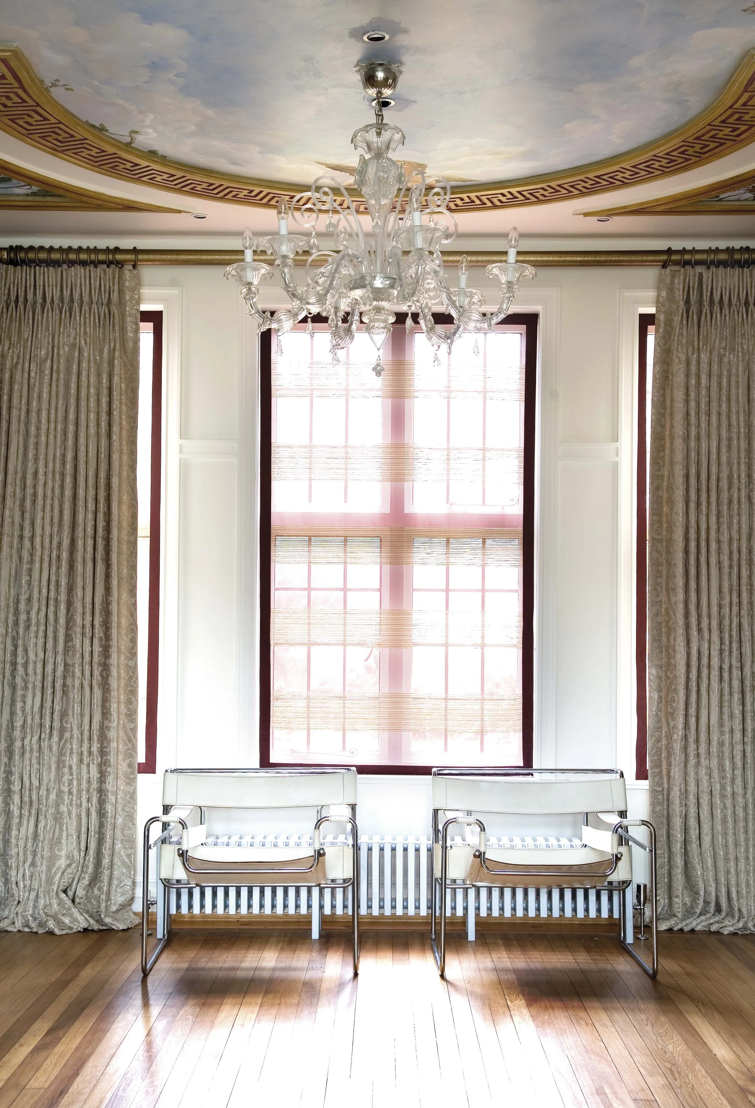 A bright room with a large window covered by a pink and beige Roman shade, beige patterned curtains on either side, a white chandelier hanging from the ceiling, and two white modern chairs with chrome frames placed against the wall below the window. The room has a painted ceiling with cloud-like details and decorative molding around the ceiling perimeter. Wooden flooring finish the space.
