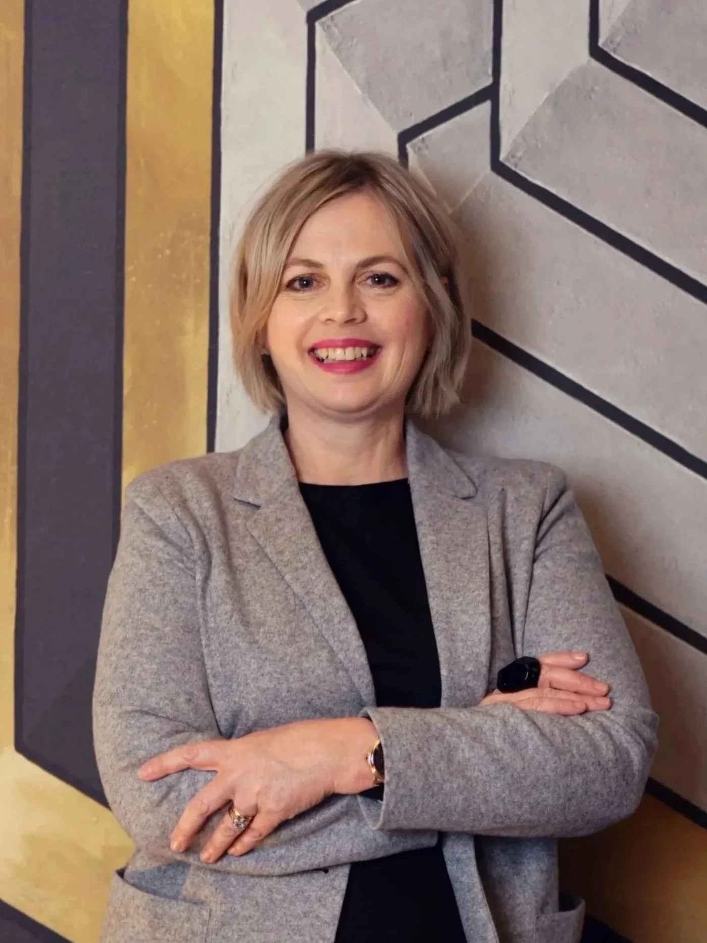 A woman with short blond hair wearing a gray blazer and black shirt, standing with arms crossed in front of a geometric patterned wall.