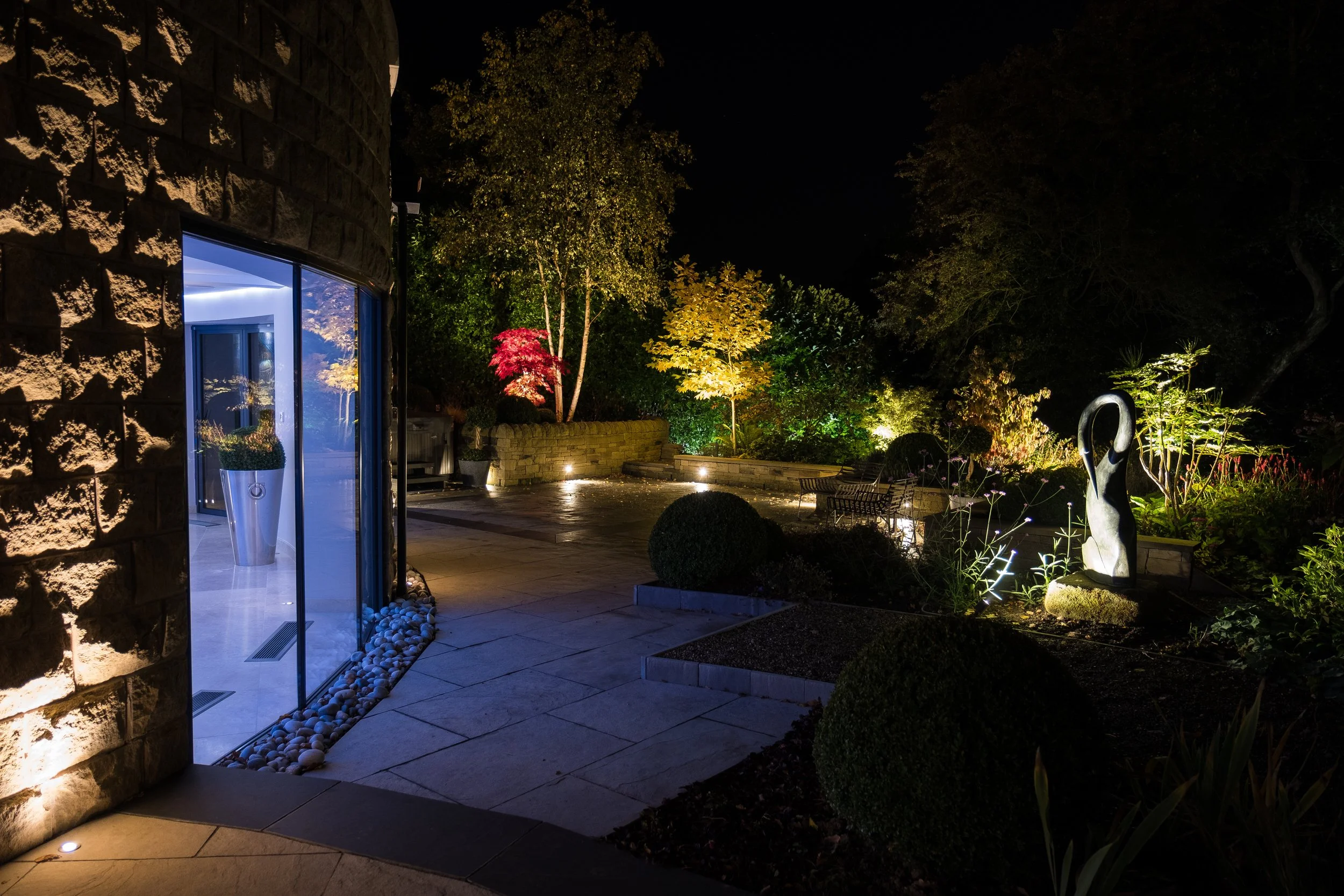 Night view of a landscaped garden with trees lit by spotlights, outdoor seating, and modern sculptures, seen from the patio of a house with lit interior, stone walls, and glass door.