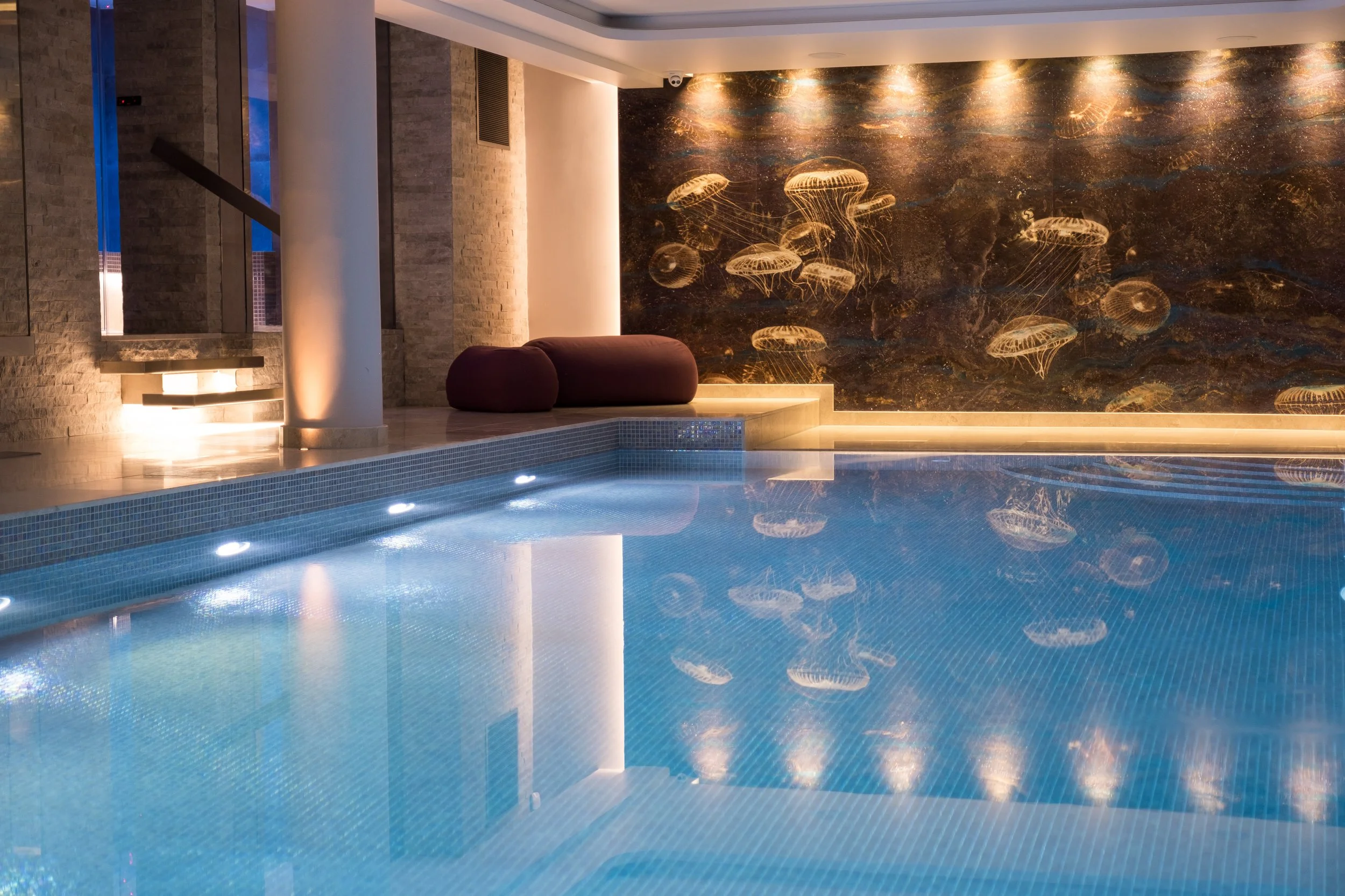 Indoor swimming pool with underwater lighting, a jellyfish-themed mural, and modern lounge seating with a stone wall and large window