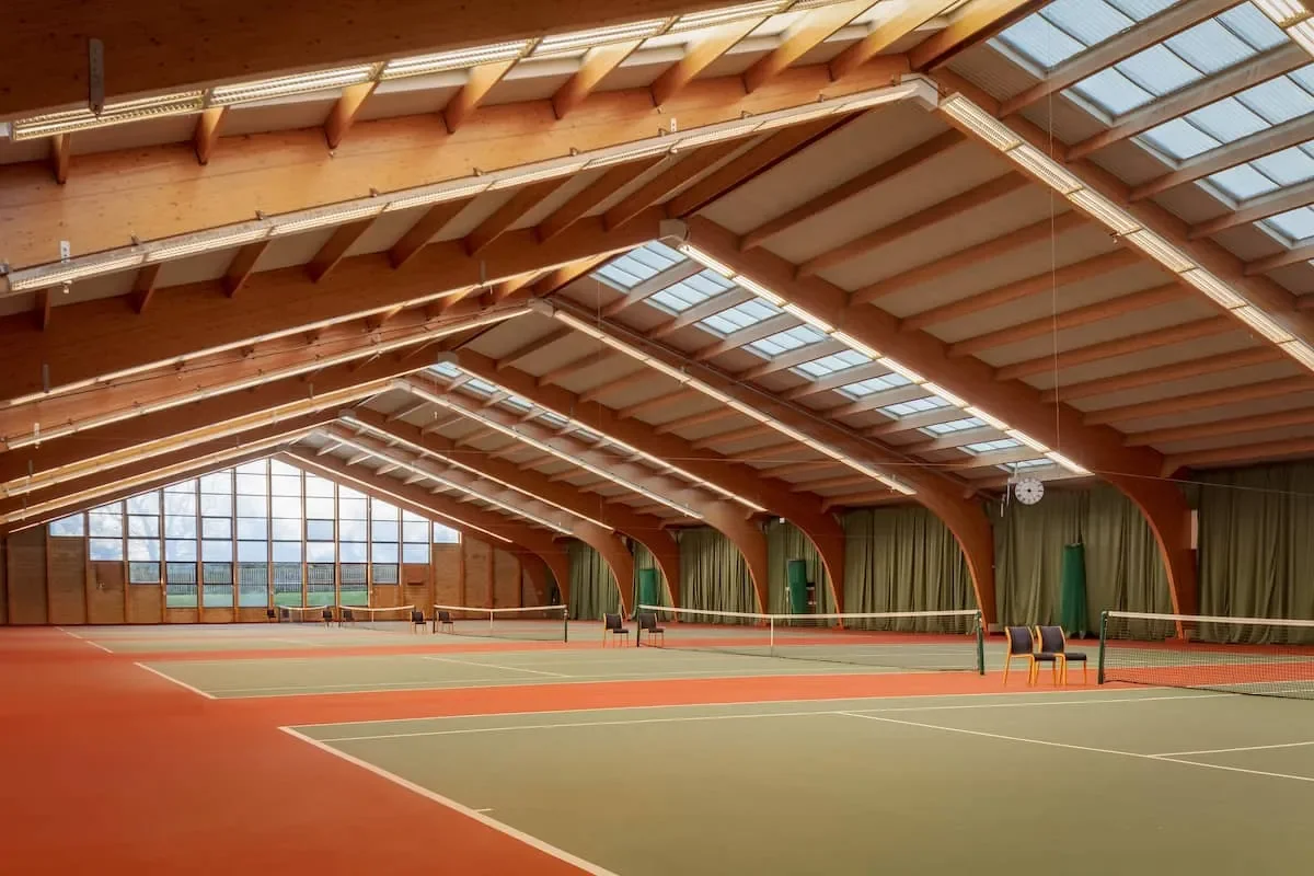 Indoor tennis courts with wooden ceiling, large windows, green curtains, and a clock on the wall.