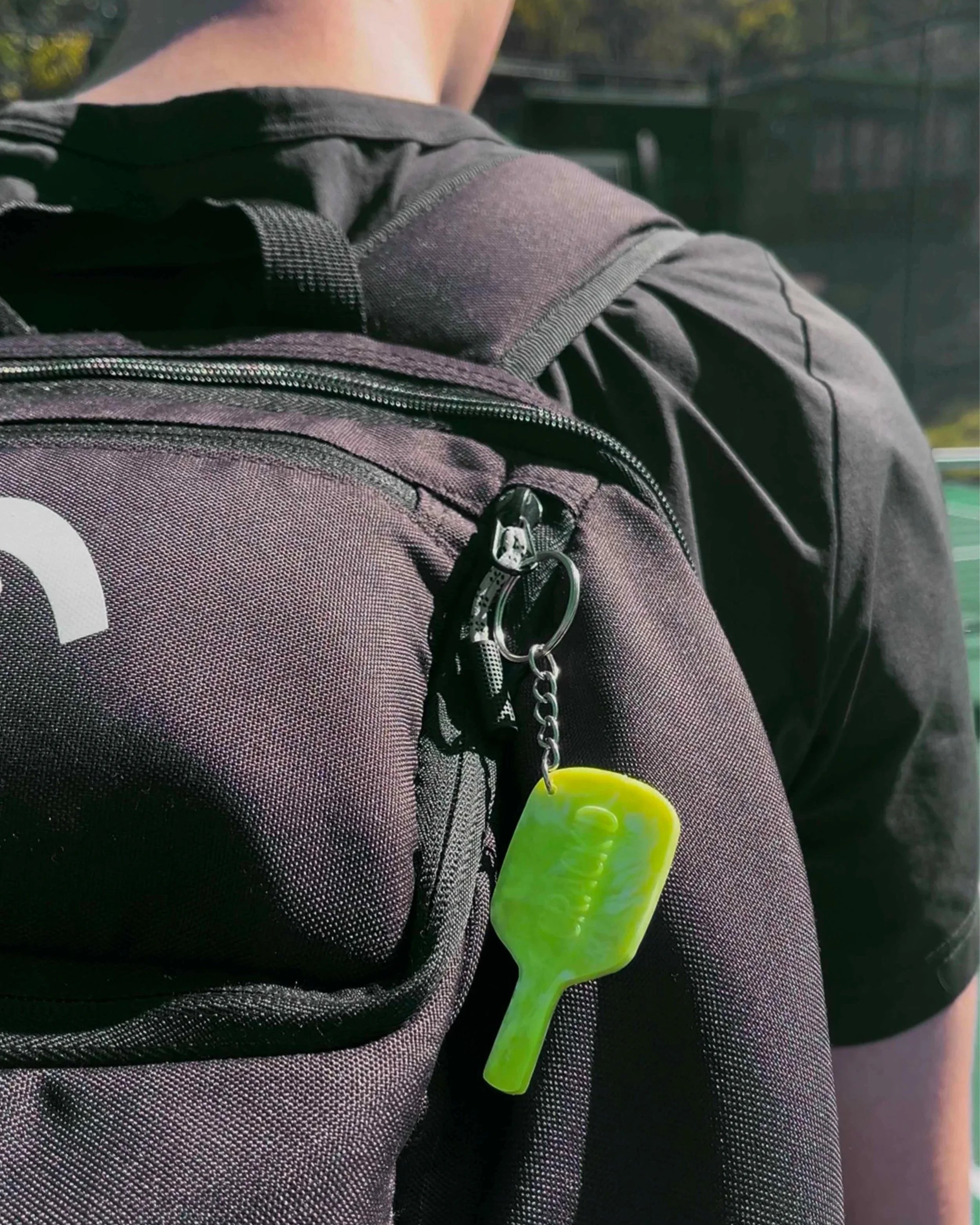 Close-up of a black and gray backpack with a zipper, attached to a person's black athletic shirt with a neon green plastic fork hanging from it.