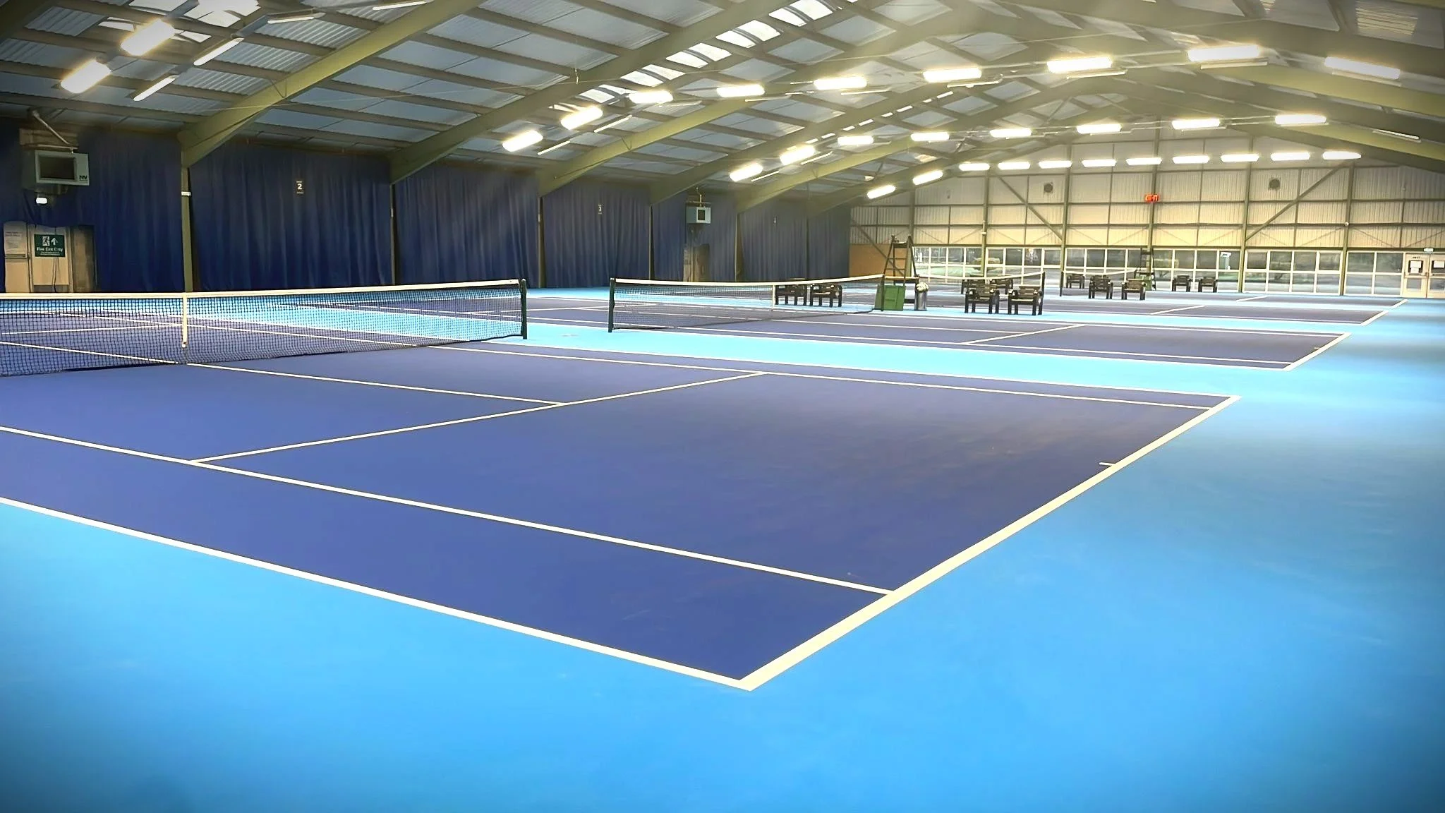 An indoor tennis court with blue surface, a net in the middle, and chairs on the side. The ceiling has lights and metal beams, with blue curtains along the walls.