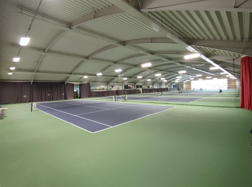 Indoor tennis courts with black courts, green surrounding flooring, and bright overhead lighting.