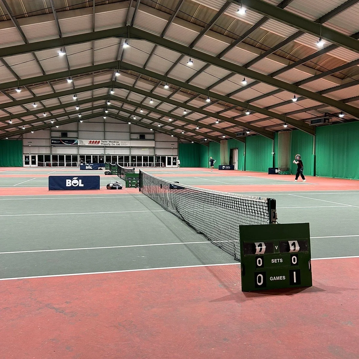An indoor tennis court with a net in the middle, surrounded by green curtains and illuminated by overhead lights. There are two players practicing, one on each side of the net, and a scoreboard showing 7-1 in favor of the second player with no sets or games completed.