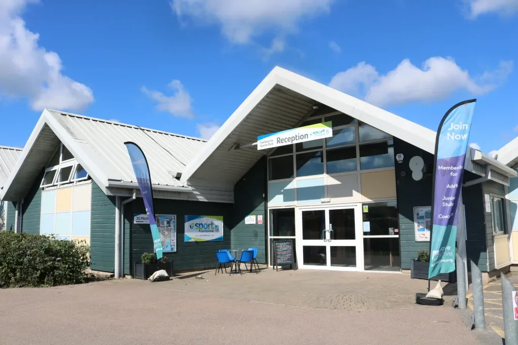 Building with signs indicating it's a sports leisure center, featuring flags with promotional text, a reception area, and outdoor seating.