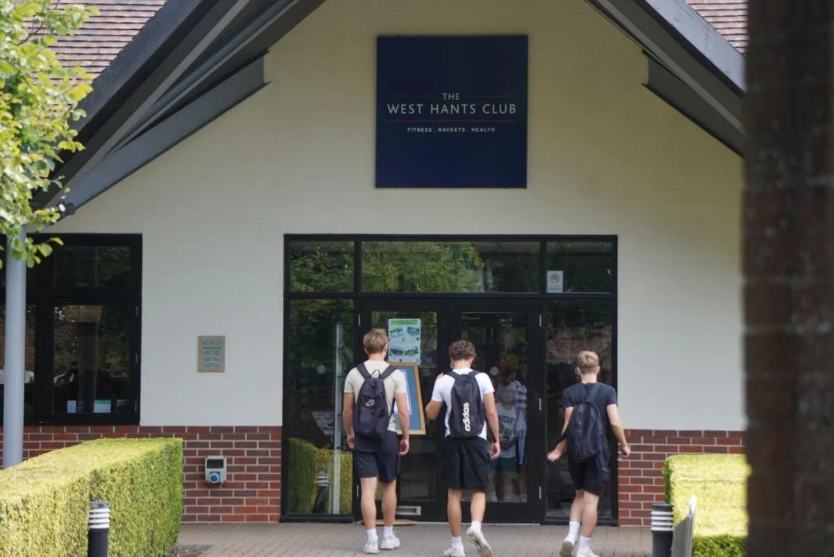 Three boys walking towards the entrance of The West Hants Club, a fitness and health center, with a sign above the door and greenery on both sides.