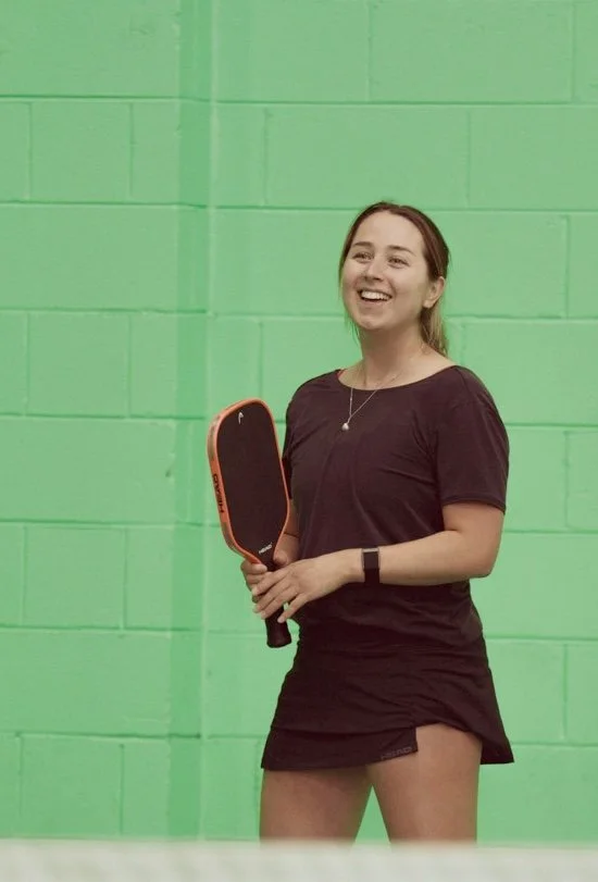 Young woman smiling and holding a pickleball paddle inside a gym with a green wall.