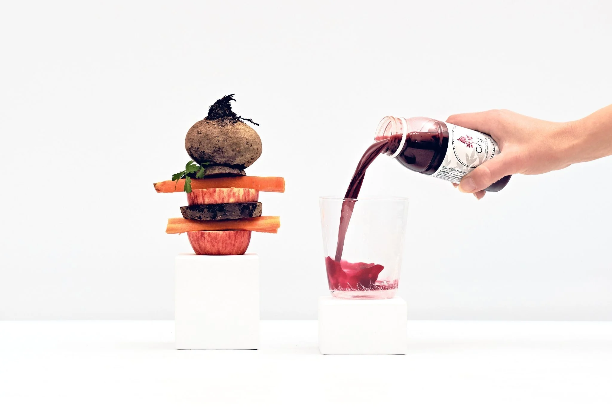 Eine Hand, die roten Gemüse-Saft in ein Glas gießt, neben einer kunstvoll gestapelten Mischung aus rotem Apfel, Karotte, schwarzer Rübe, Rote Beete, und einem Radieschen, alles auf weißen kubischen Ständern