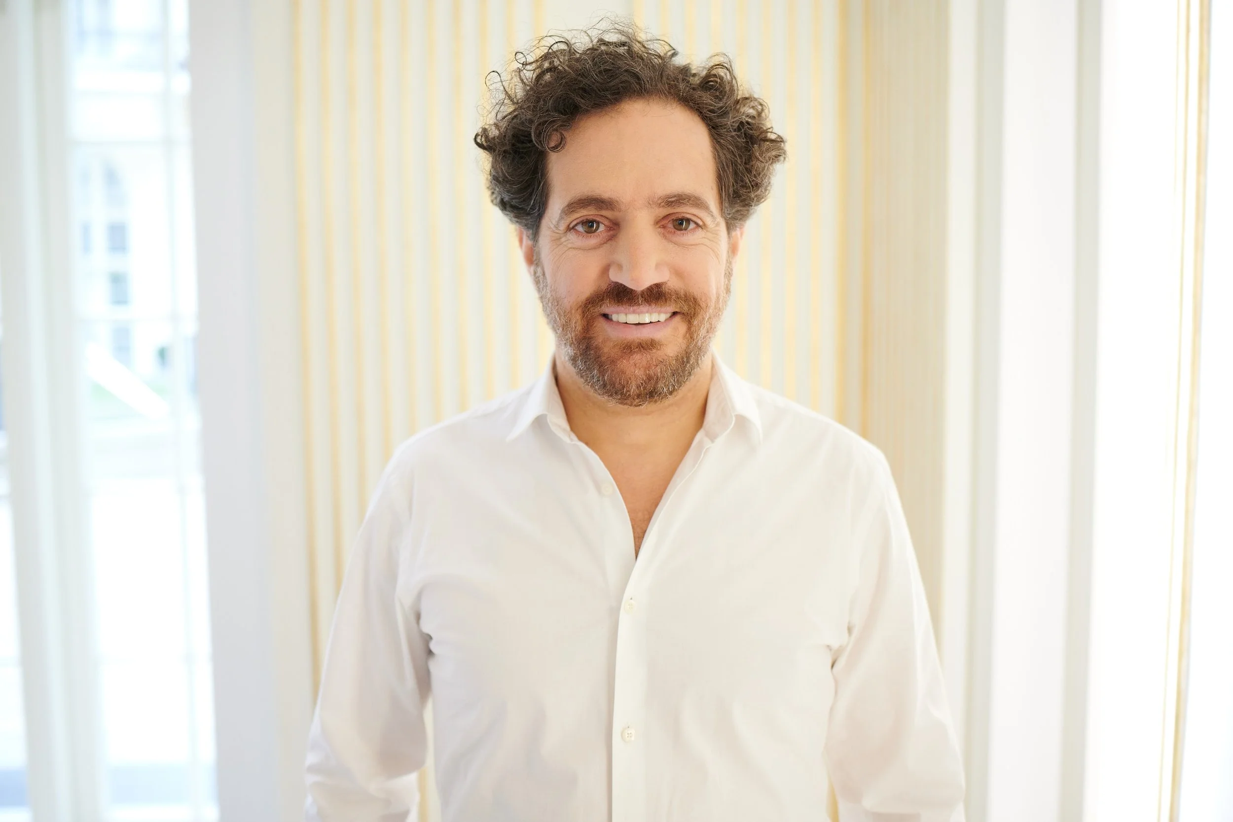 Ein Mann mit braunem, lockigem Haar und Bart, trägt ein weißes Hemd, steht vor einem Fenster mit gelb-gestreiftem Vorhang, lächelt in die Kamera.