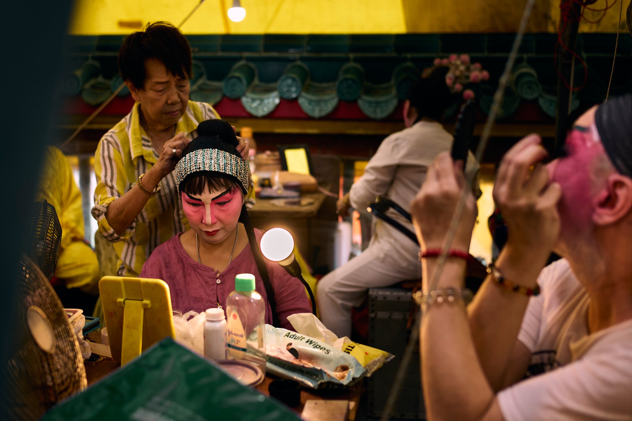 A backstage scene showing a Chinese opera performer having hair done before a performance, surrounded by mirrors, lights, and fellow performers in preparation.