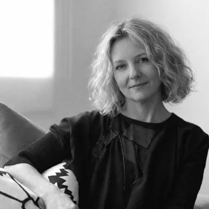 A woman with curly hair sitting on a sofa in a room with natural light, smiling softly at the camera.