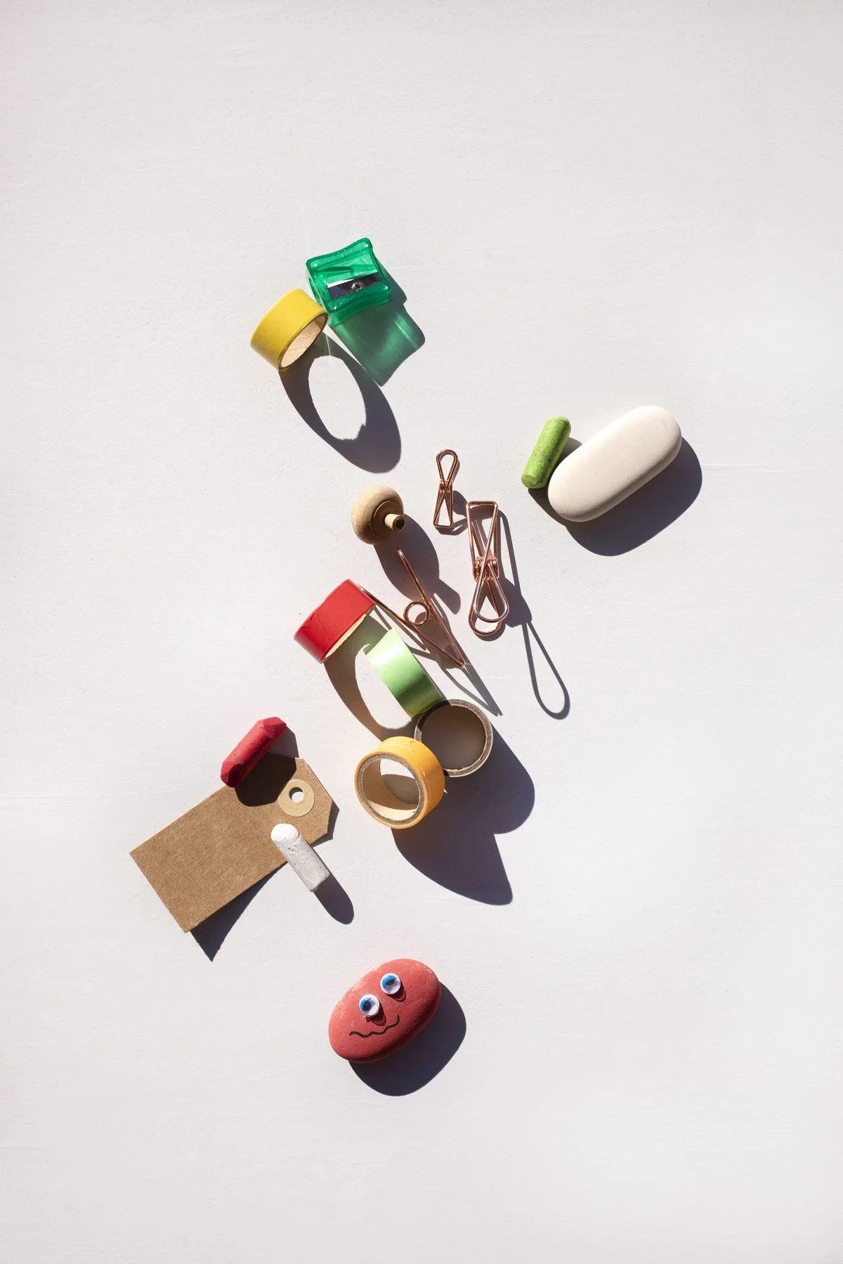 Conjunto de cinta adhesiva de colores, clips de papel, una piedra decorativa con caras y otros objetos pequeños dispuestos en una superficie blanca con sombra.