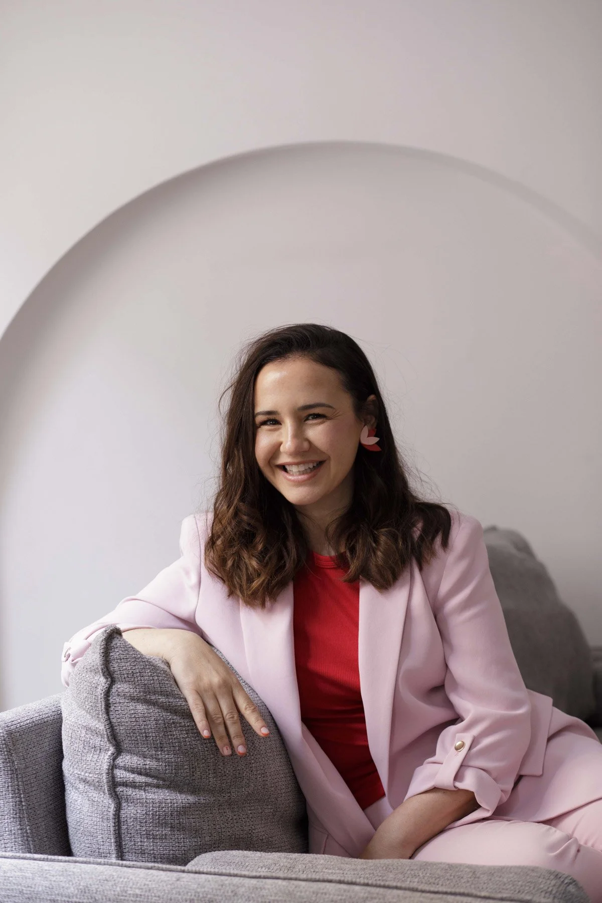 Lidia Viso sonriendo, sentada en un sillón gris, vestida con blazer rosa y camiseta roja, con pendientes color rosa y blanco.