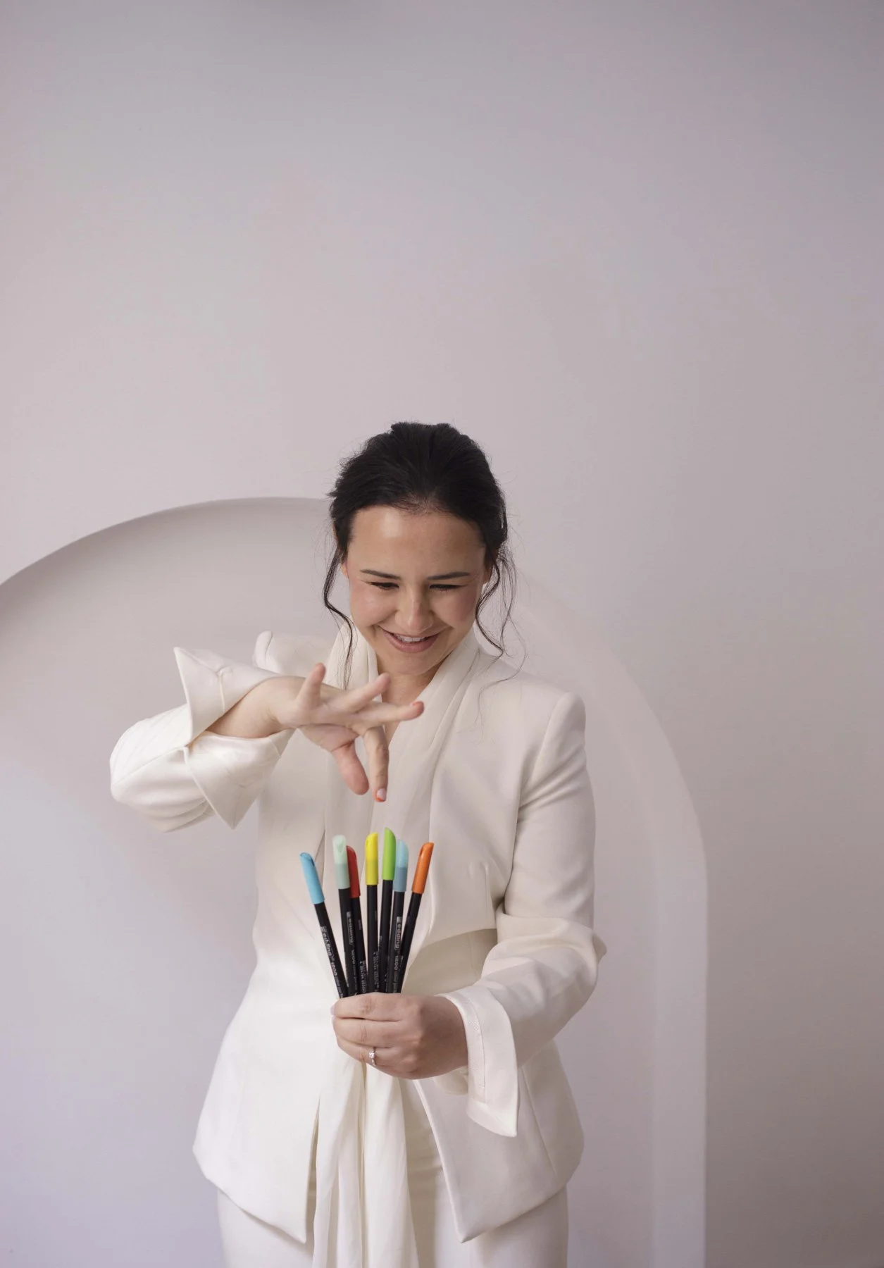 Lidia Viso sonriendo con cabello oscuro, vestida con traje blanco, sosteniendo varios rotuladores de colores, en un fondo blanco con un arco decorativo
